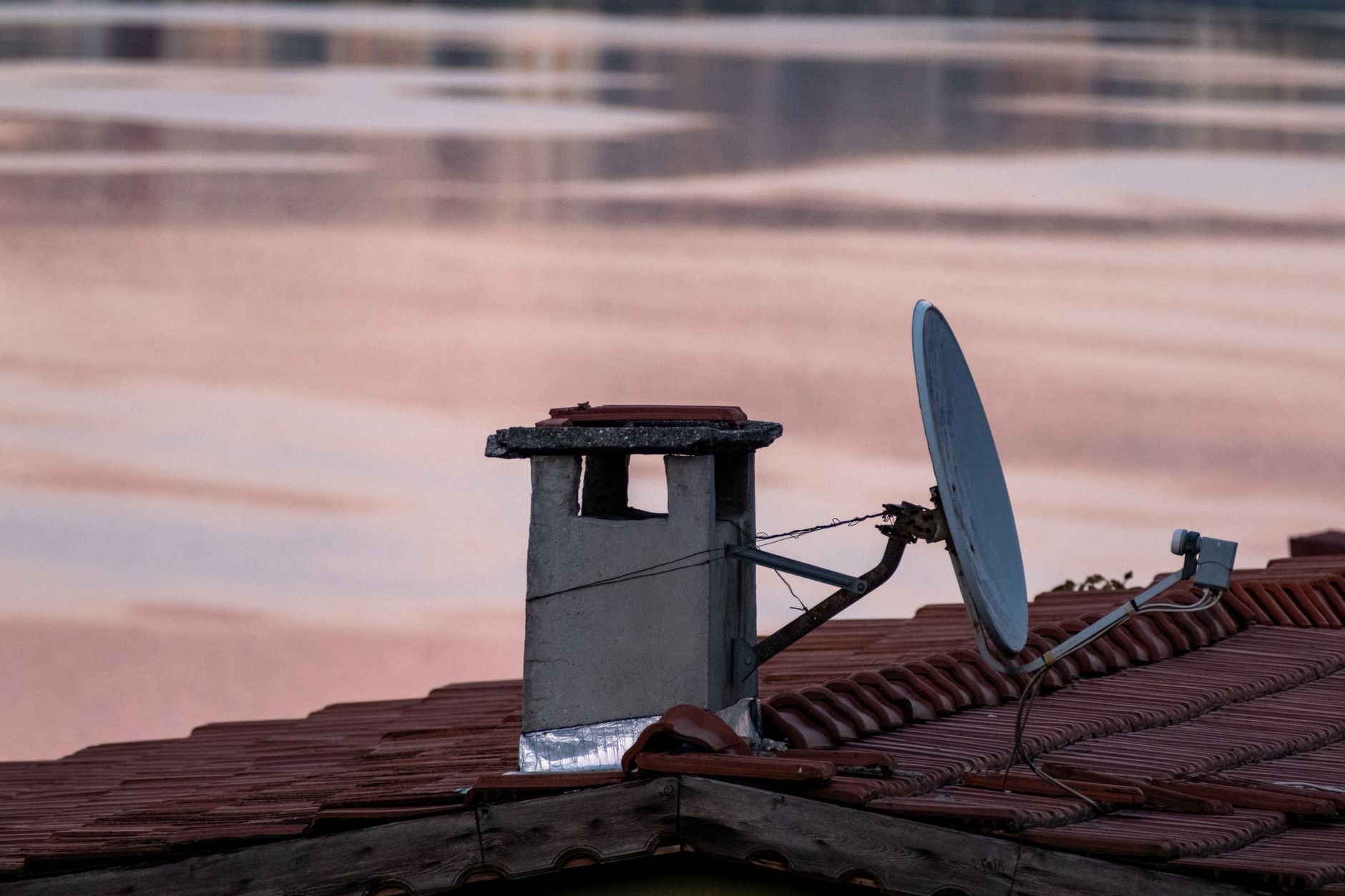 Grey satellite dish on a red-tiled roof beside a concrete chimney, with a pink-tinted lake or water behind