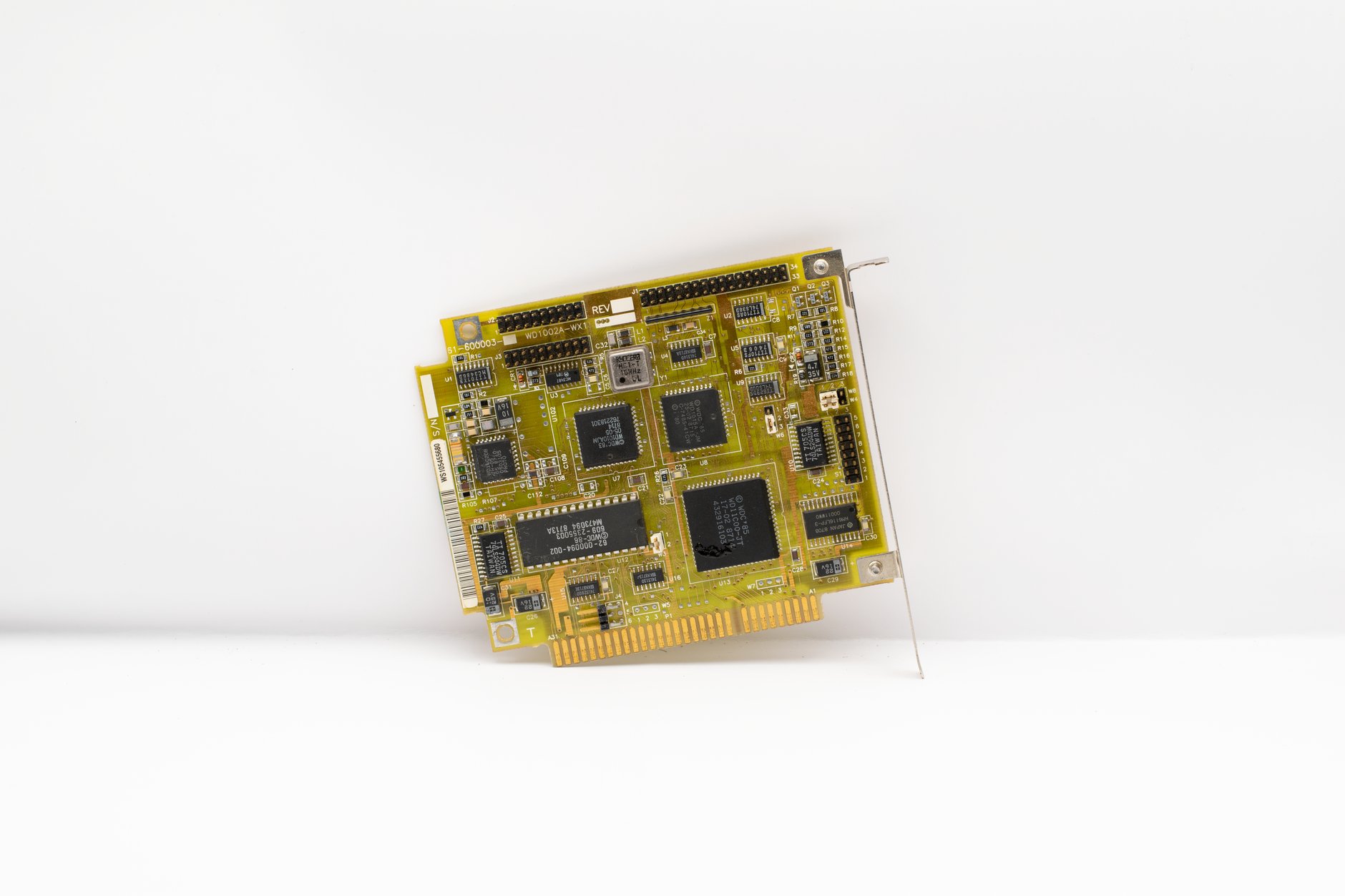 Yellow circuit board with black microchips, connector pins and a metal mounting bracket against a plain white background