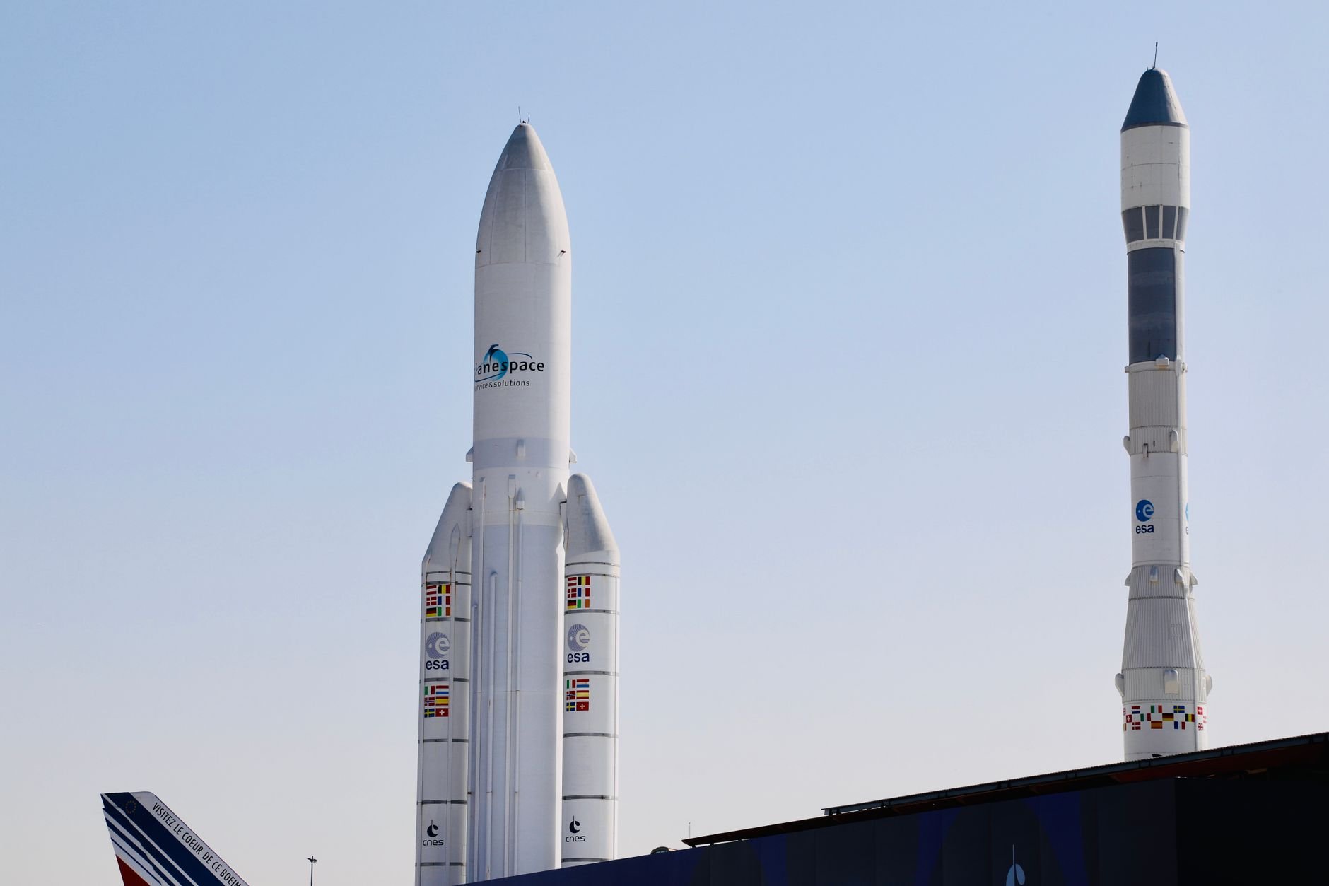 Two tall white launch vehicles standing upright under a clear blue sky, with Arianespace branding visible on one