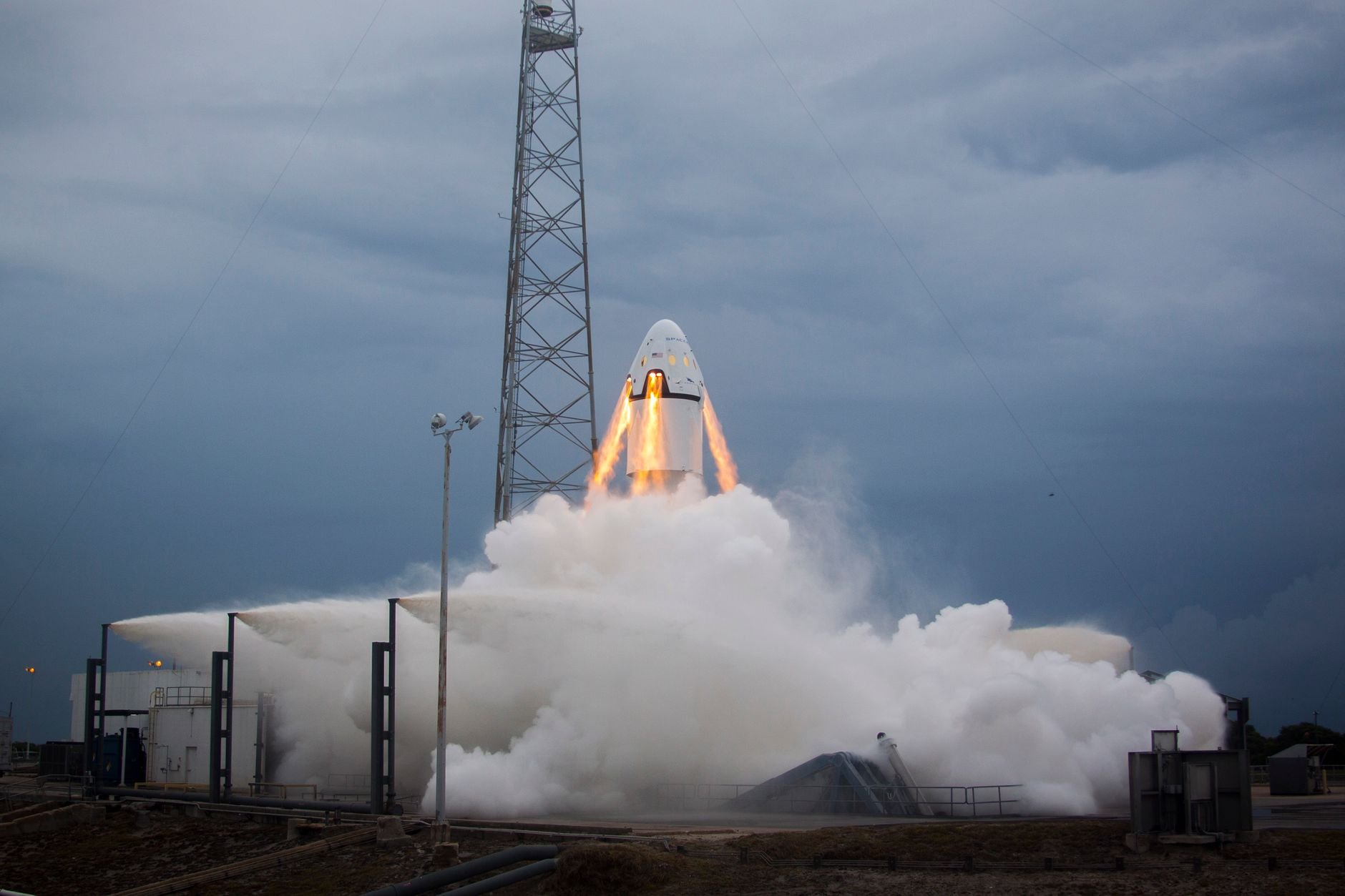 Capsule-shaped spacecraft with engine flames at the base, surrounded by a large cloud of vapour at a launch pad under a stormy sky