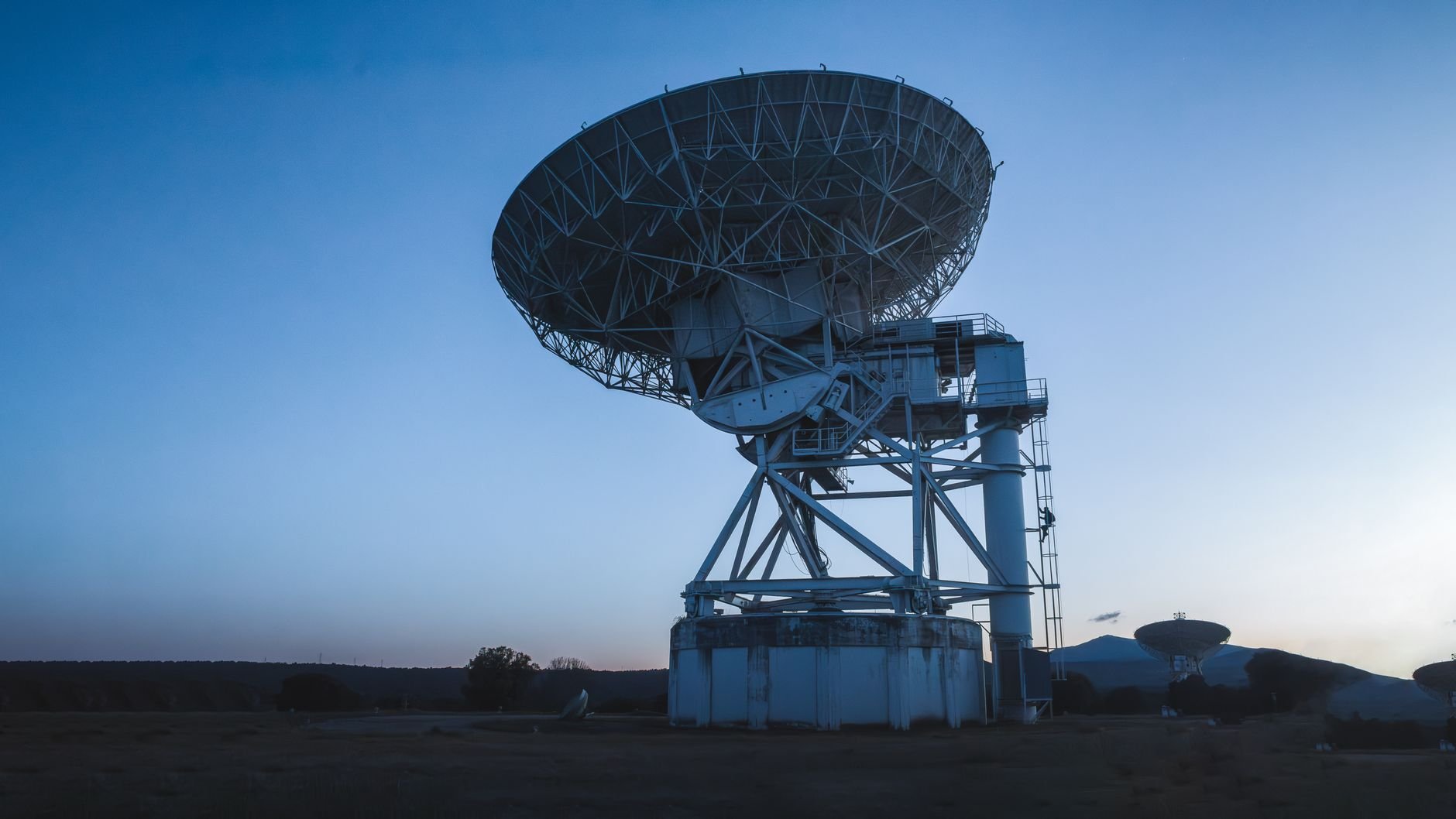 Large radio dish antenna on a steel mount under a deep blue dusk sky, with smaller dishes in the distance