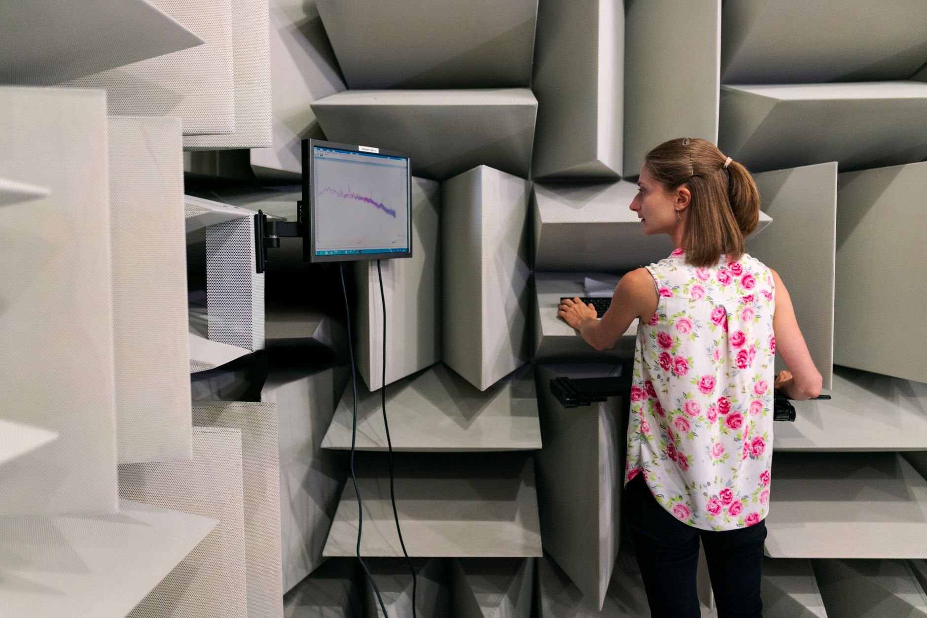 Woman at a monitor inside an anechoic chamber whose walls are covered in grey foam absorber wedges