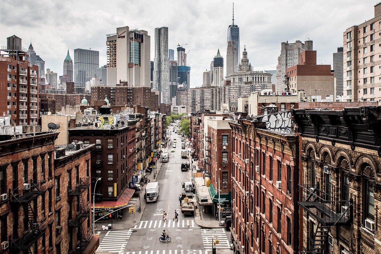 Elevated view of a Manhattan-style street canyon with brick and fire-escape apartment buildings and tall skyscrapers under an overcast sky