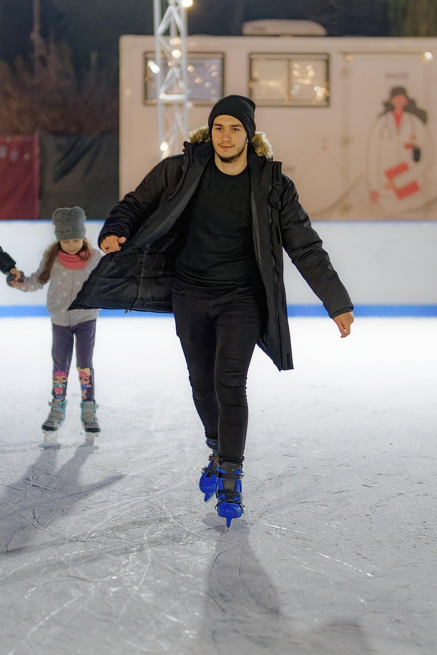 Man in a dark hooded jacket and black clothing skates on blue-bladed skates on an outdoor rink at night, with a young girl in a grey hat skating behind him