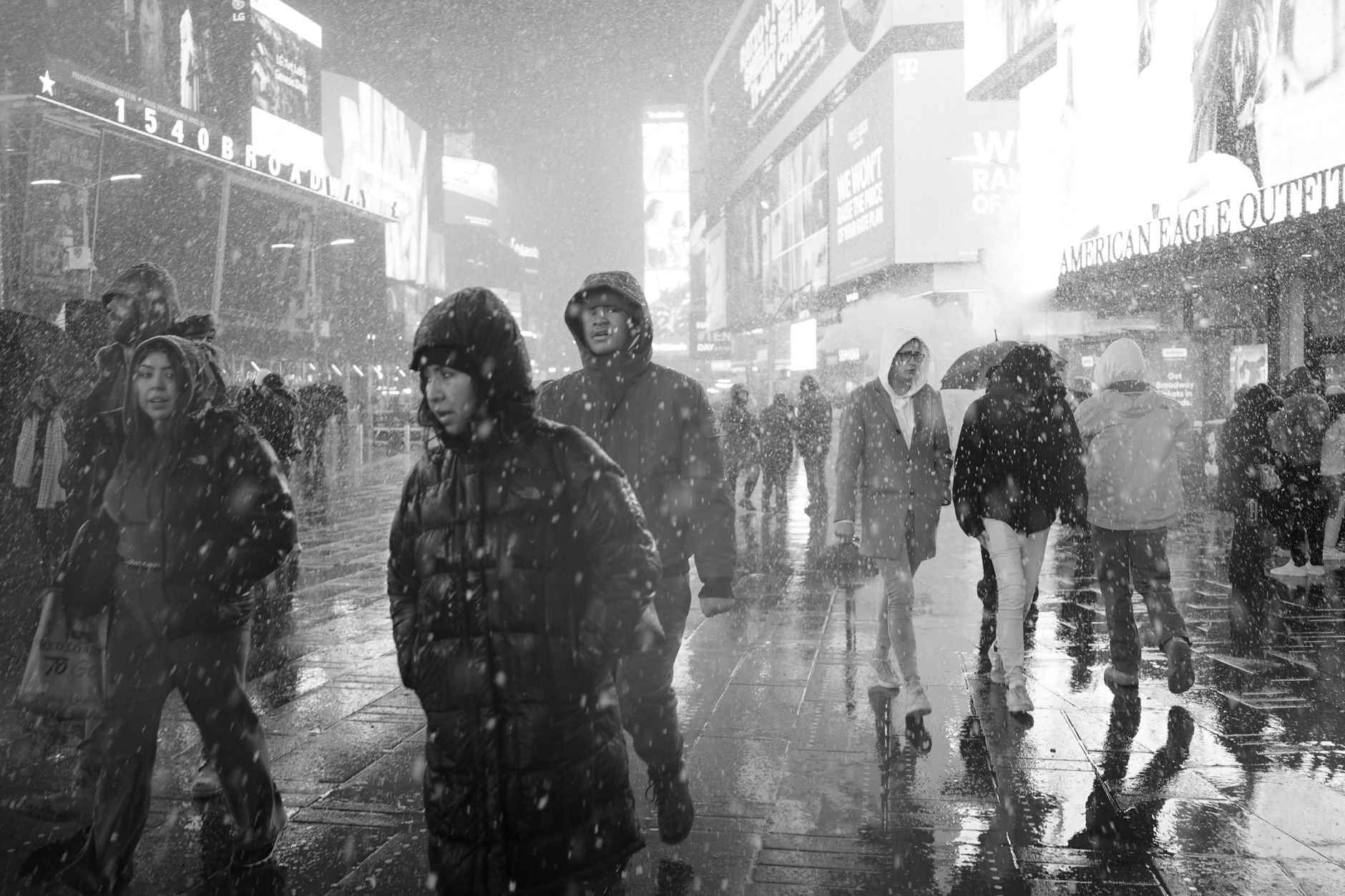 Winter crowds in heavy hooded coats walking on a snowy, wet city sidewalk at night in black and white