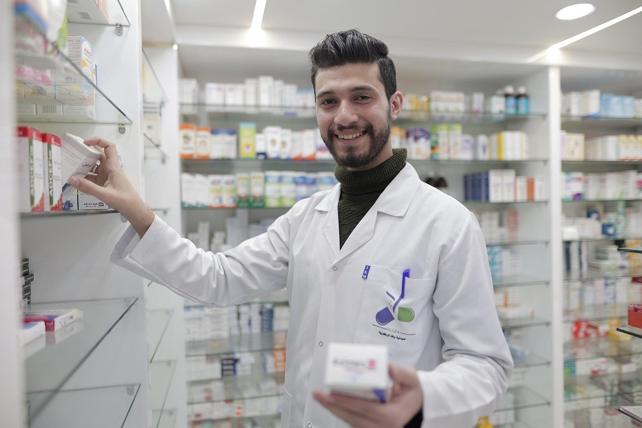 A smiling bearded man in a white lab coat stands in a pharmacy, reaching toward shelves of boxed products and holding a small white box.