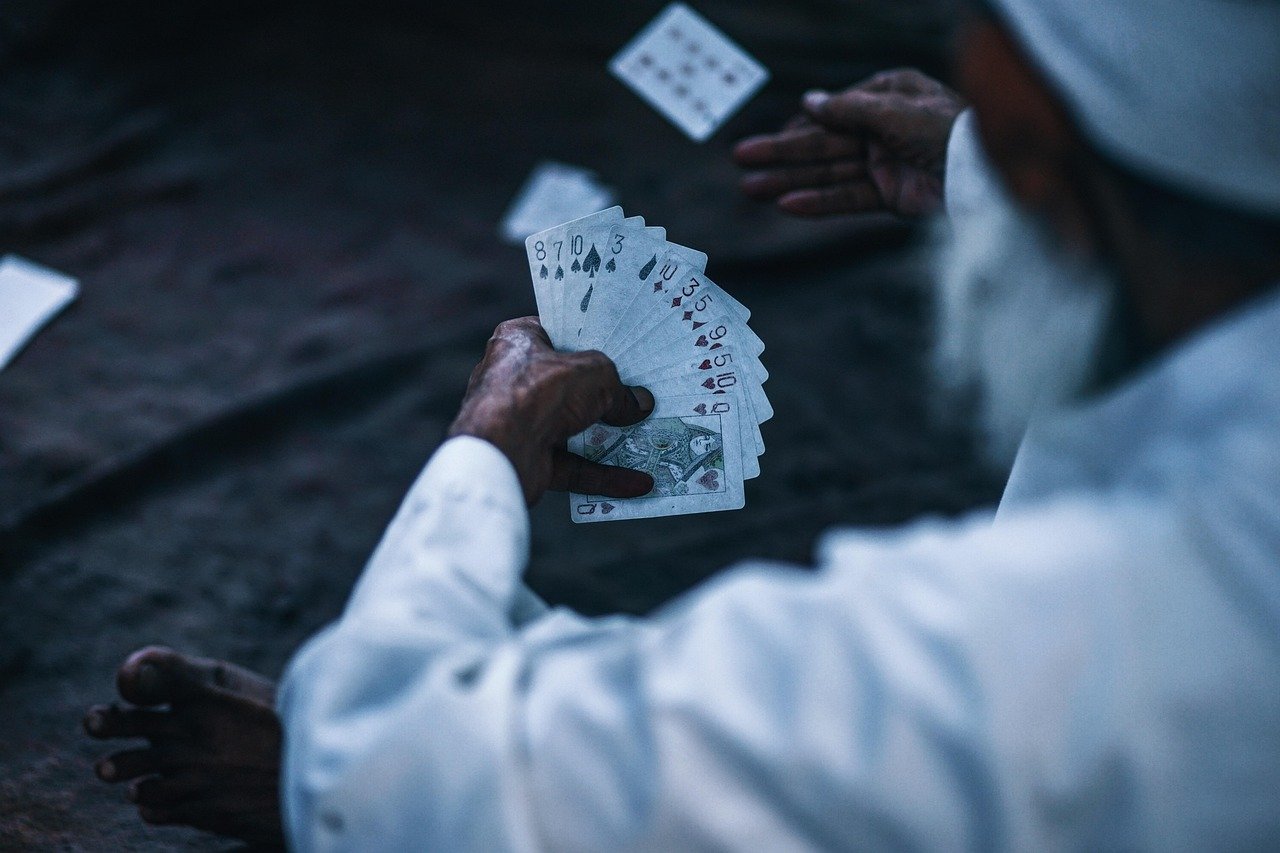 An elderly bearded man in a white robe holds a fan of playing cards in one hand while another hand gestures, with a card in the air at upper left.