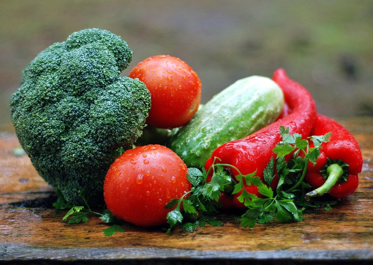 Broccoli head with two red tomatoes, a green cucumber, red bell peppers, and parsley on a wooden board, with water droplets on the produce