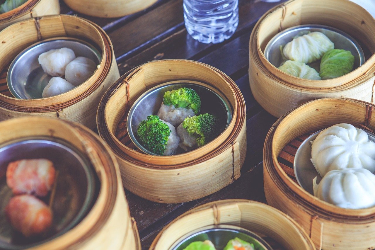 Bamboo steamer baskets of dim sum on a wooden table, including one basket with cooked broccoli florets beside dumplings
