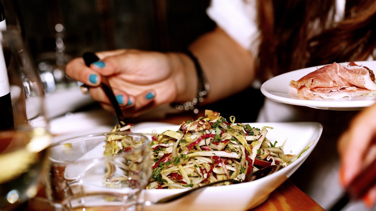 Hands holding a fork over a plate of shredded salad with red strips and herbs, beside a plate of cured meat and a glass of wine