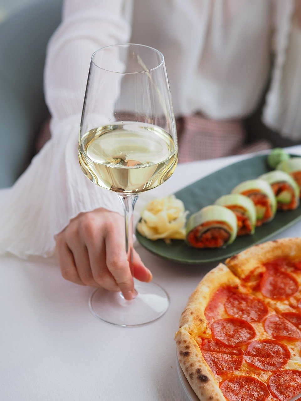Hand holding a stemmed glass of white wine above a plate of sushi rolls and a pepperoni pizza on a white table