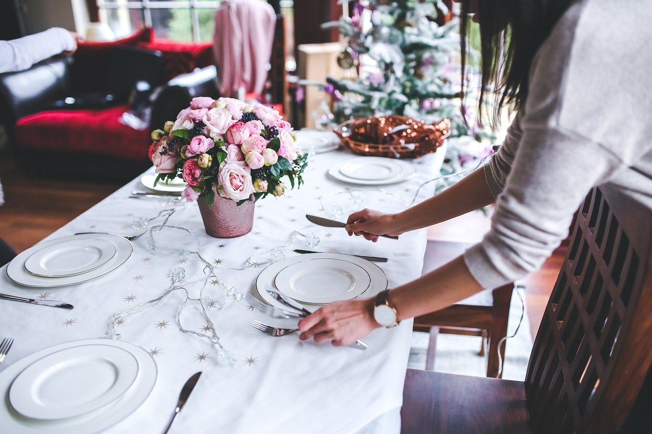 Person setting a table with white plates, silverware, and a pink flower centerpiece, with a decorated Christmas tree in the background