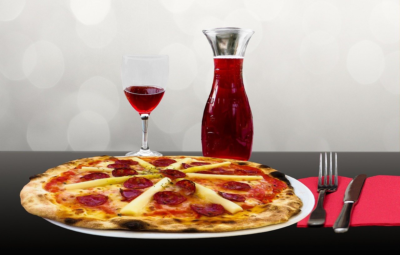 Round pepperoni pizza with pale cheese slices on a white plate beside a stemmed glass of red wine and a clear carafe of red wine on a dark table