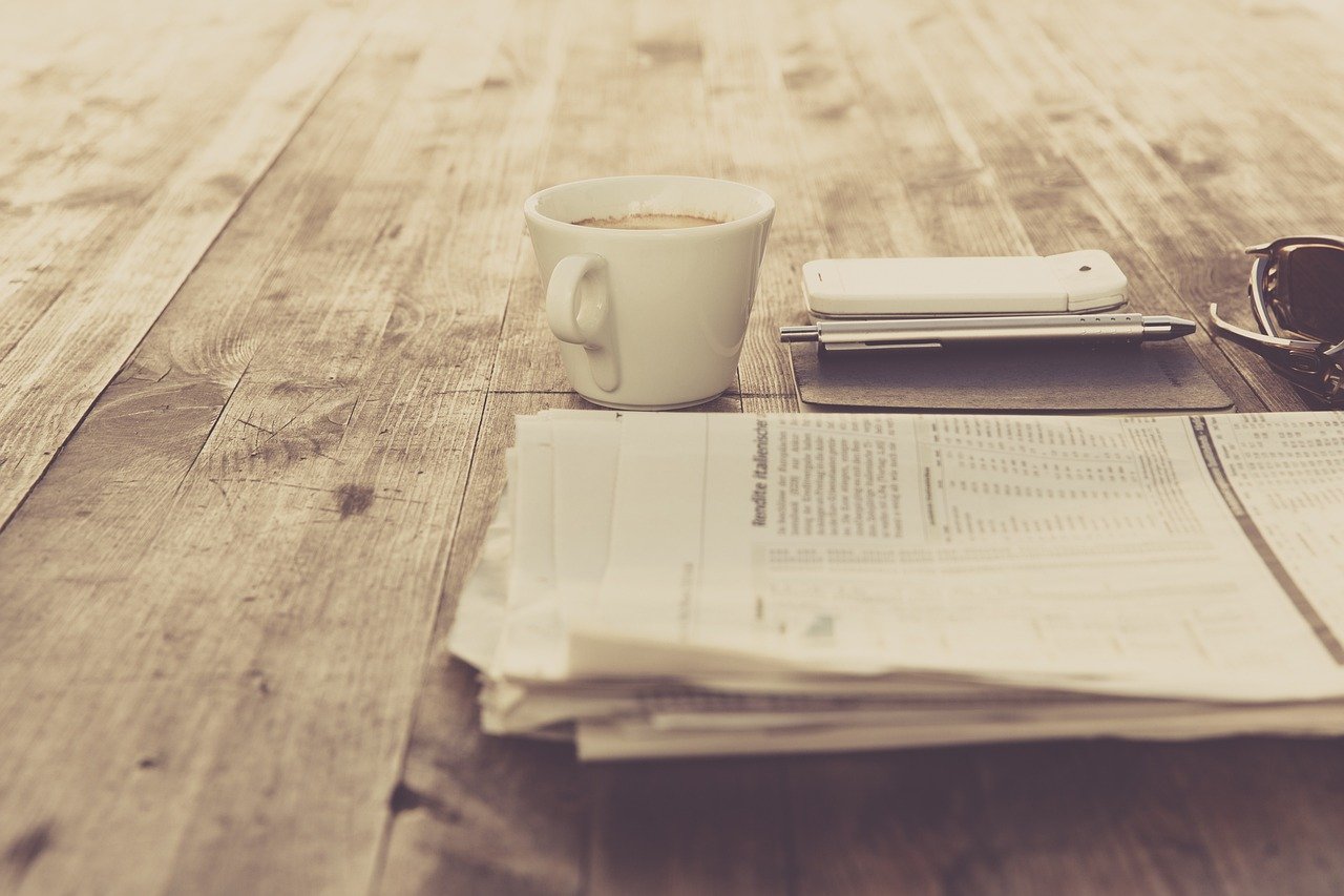 Stack of folded newspapers on a wooden table beside a cup of coffee, a phone, a pen, and sunglasses