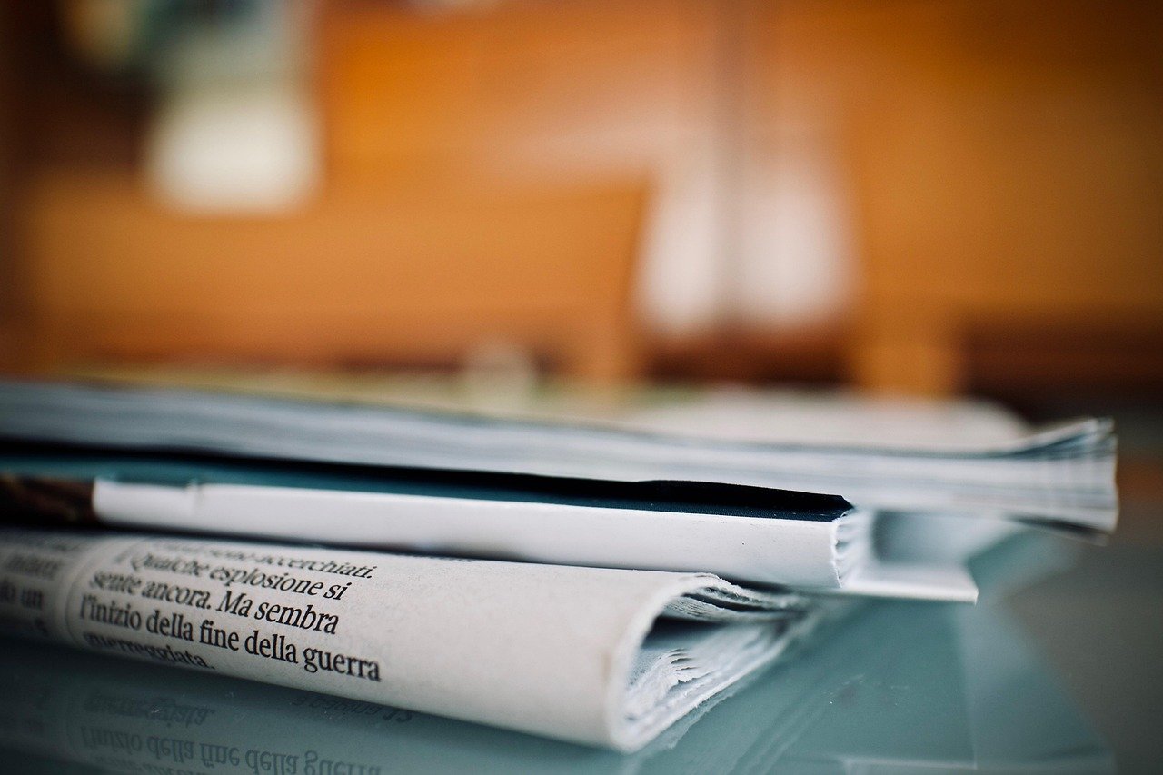 Stacked newspapers and magazines on a glass table, with Italian headline text visible on the top newspaper