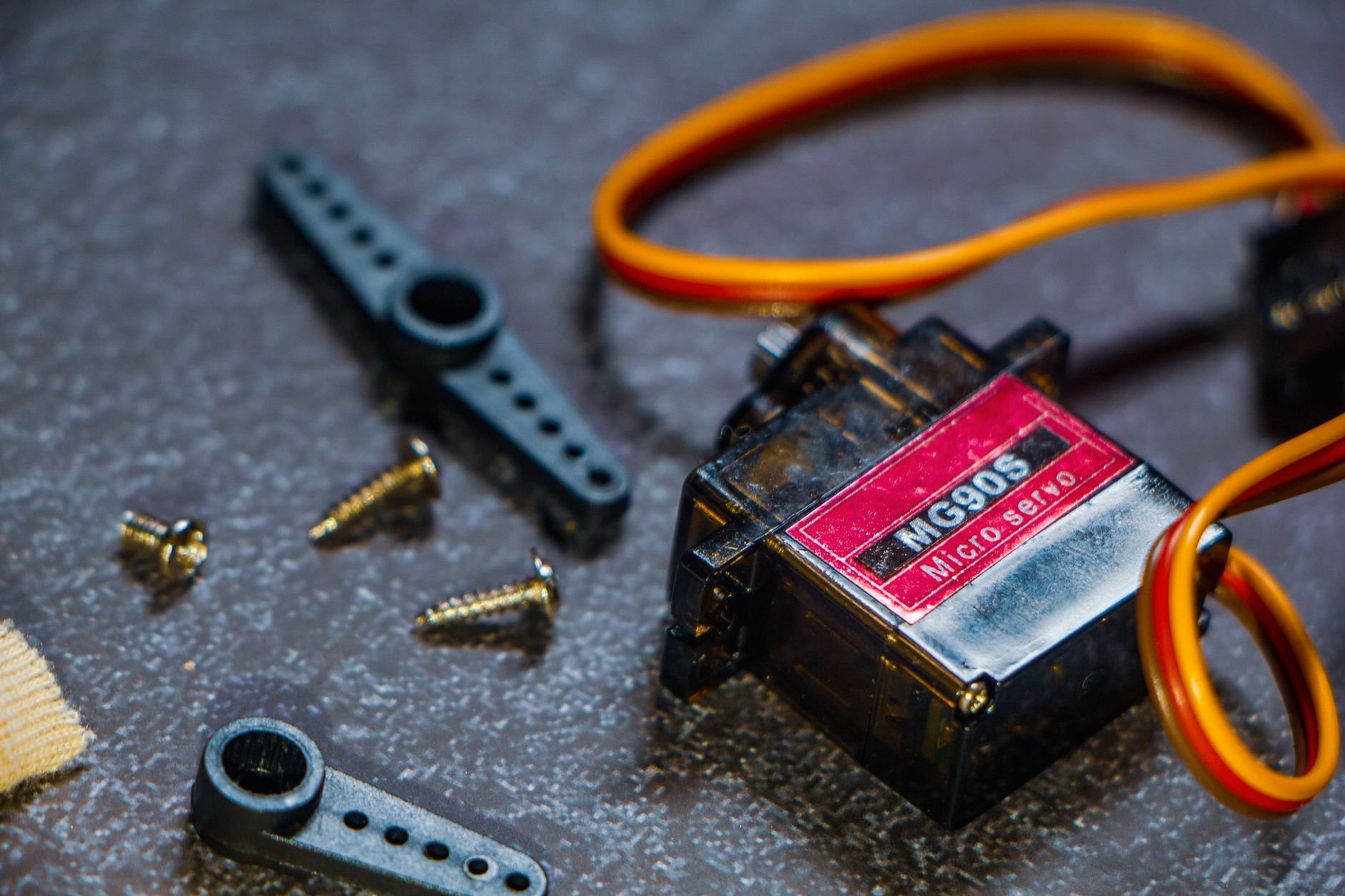 MG90S micro servo with a red label, orange wires, plastic servo horns and small screws on a dark surface
