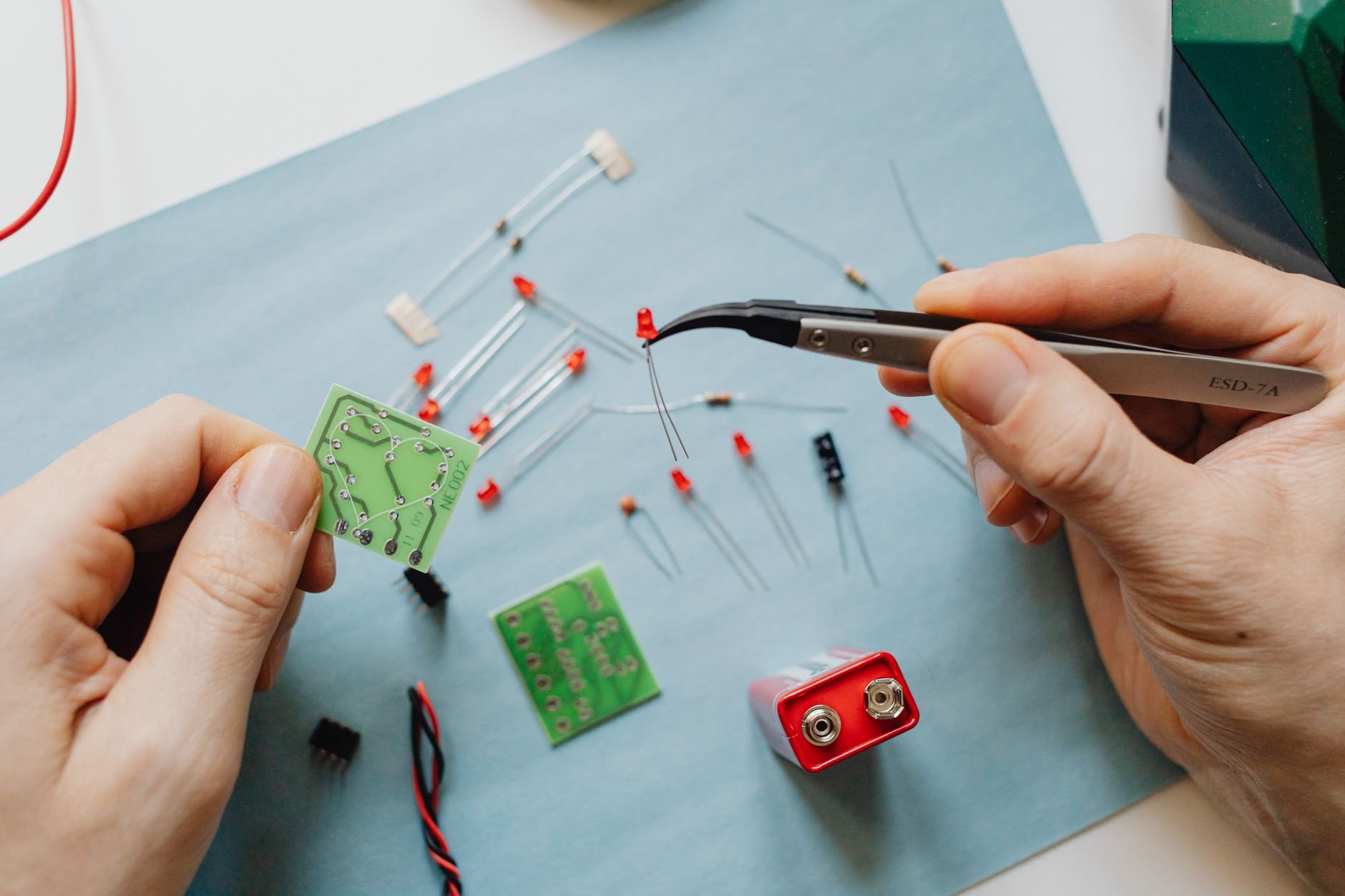 Two hands using tweezers to handle small LEDs, a battery holder and green circuit boards on a blue sheet