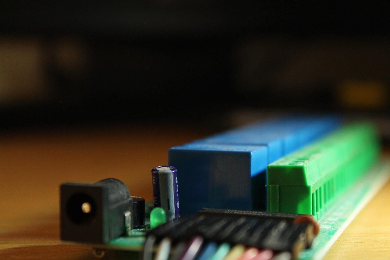 Close-up of a green circuit board edge with a black DC power jack, a blue component, a green terminal block, and a purple capacitor, against a blurred dark background