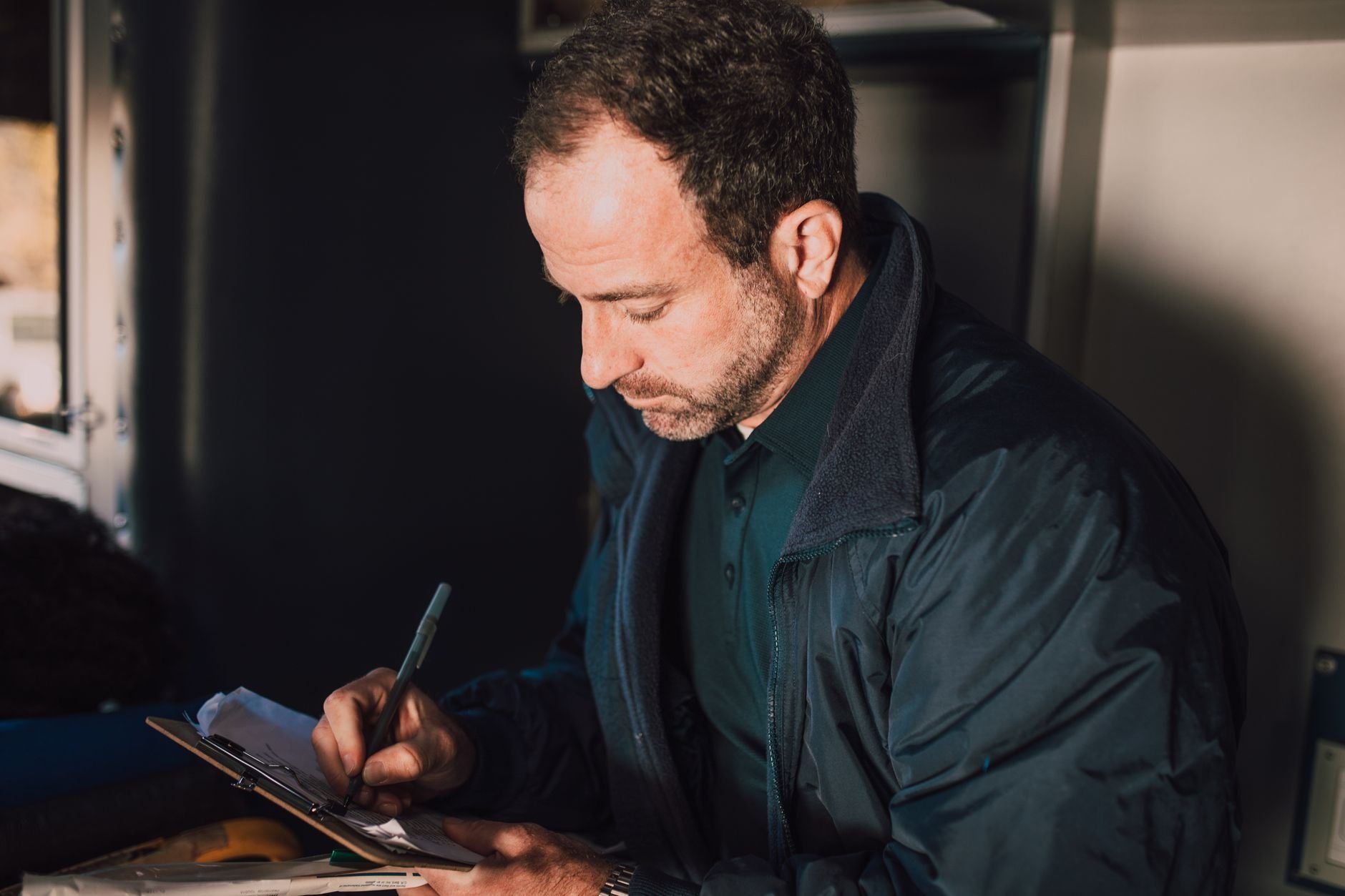 Man in a dark jacket writing on a clipboard with a pen in dim light
