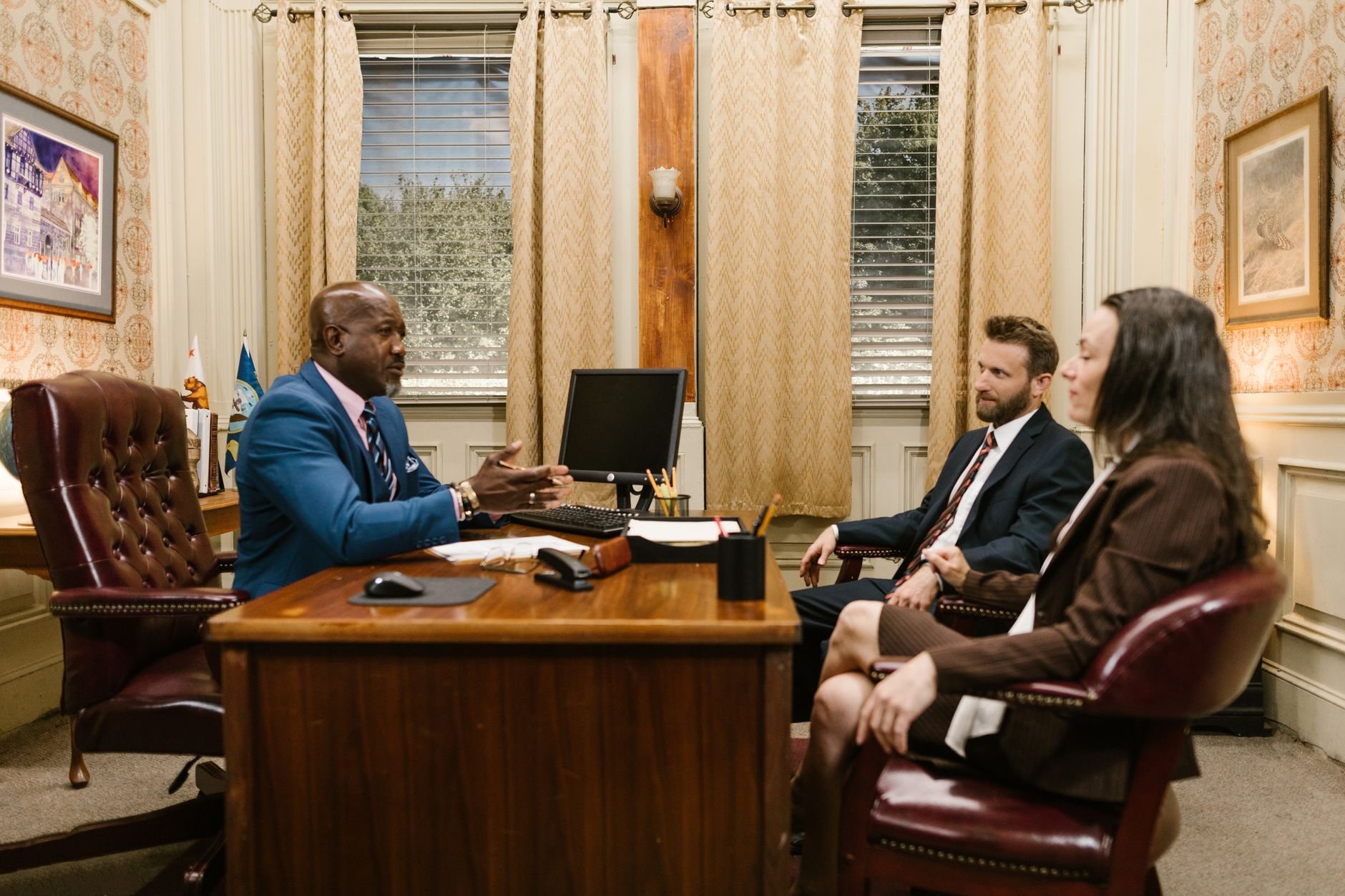 Man in a blue suit gesturing at a wooden desk while two people sit across from him in leather chairs, in a formal office with patterned wallpaper, curtains, and framed pictures.