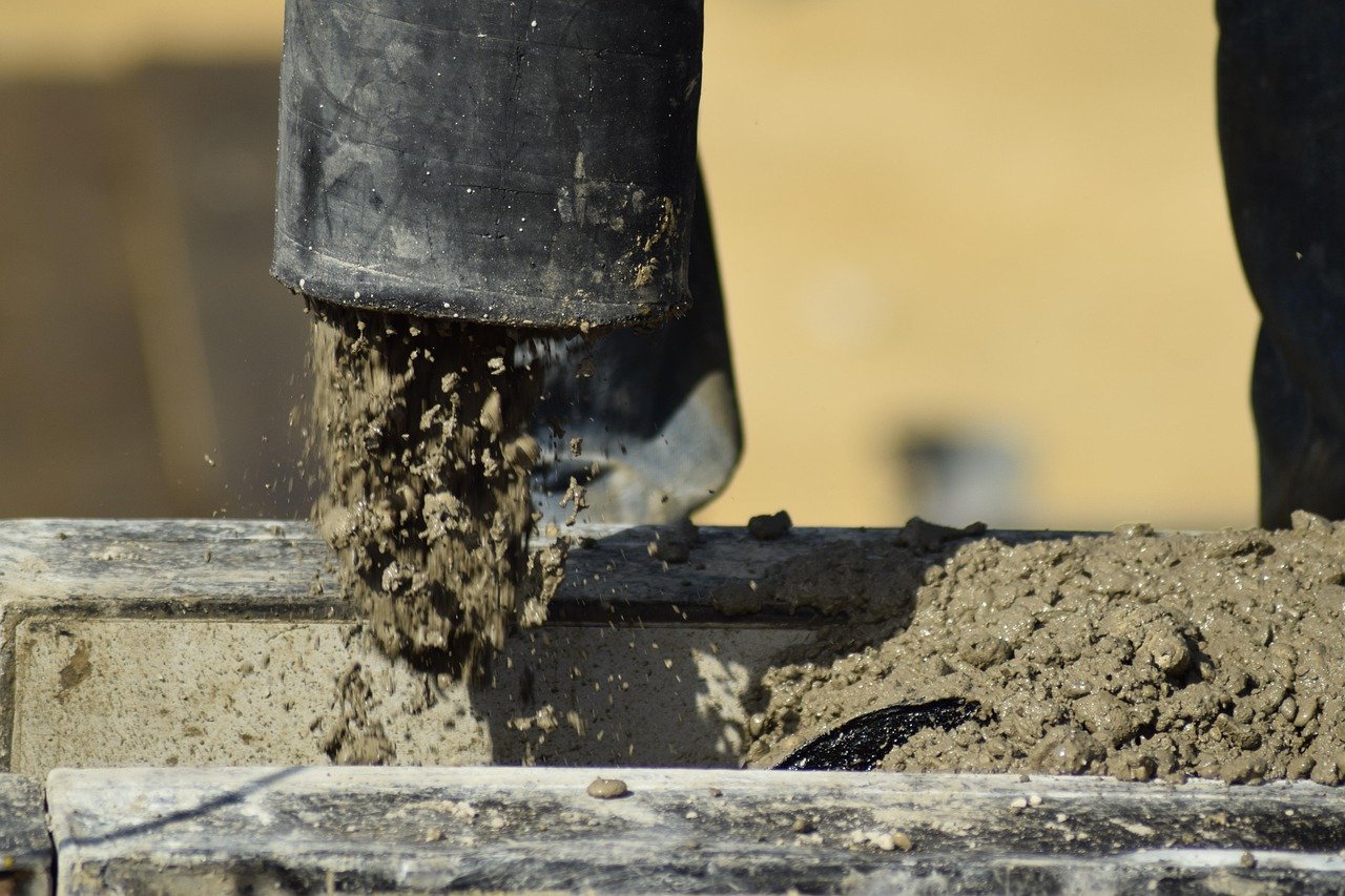 Grey concrete pouring from a black hose into a formwork edge at a construction site, with a sandy background