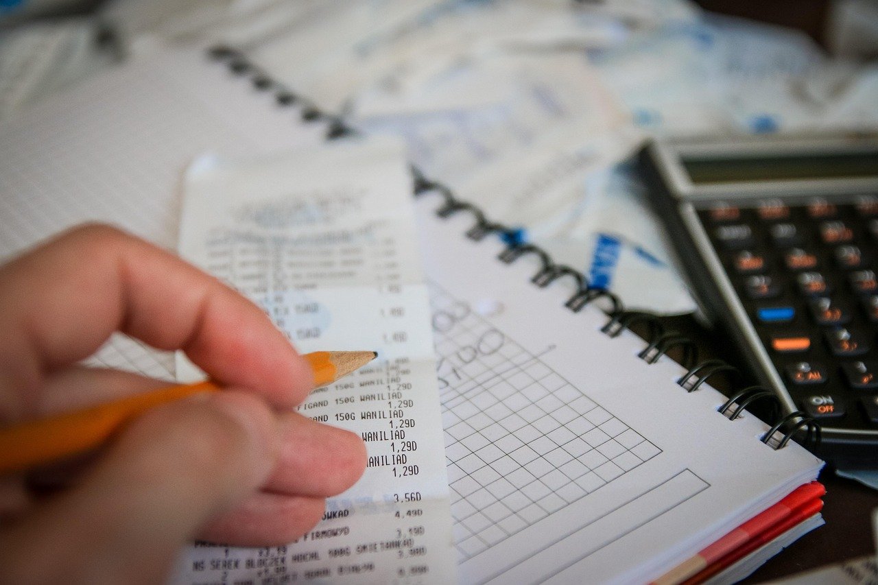 A hand holding a pencil over a paper receipt beside a spiral notebook and a desk calculator