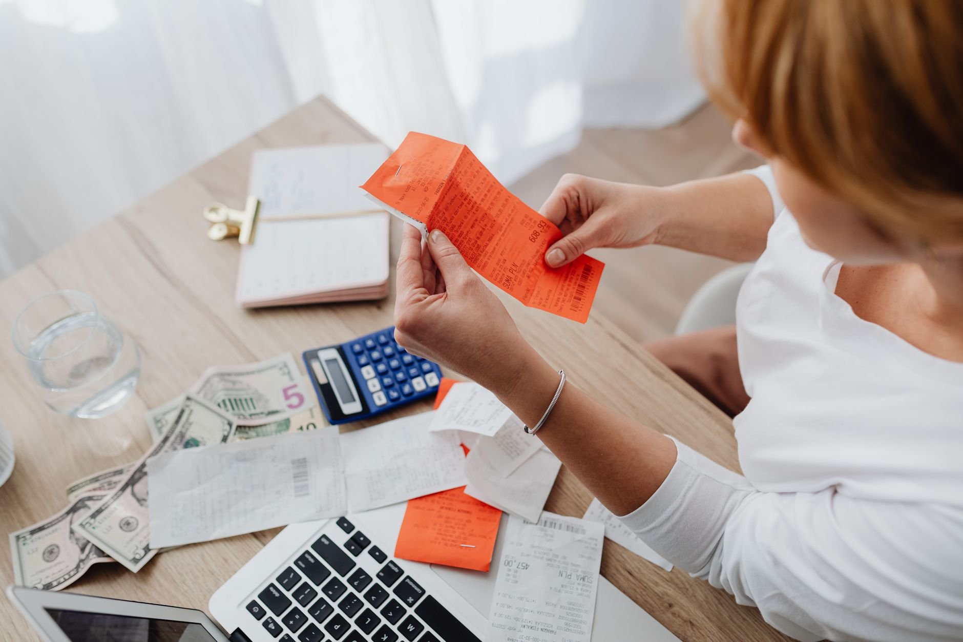 Woman at a wooden table holding an orange paper slip beside a blue calculator, loose white papers, banknotes and a laptop keyboard