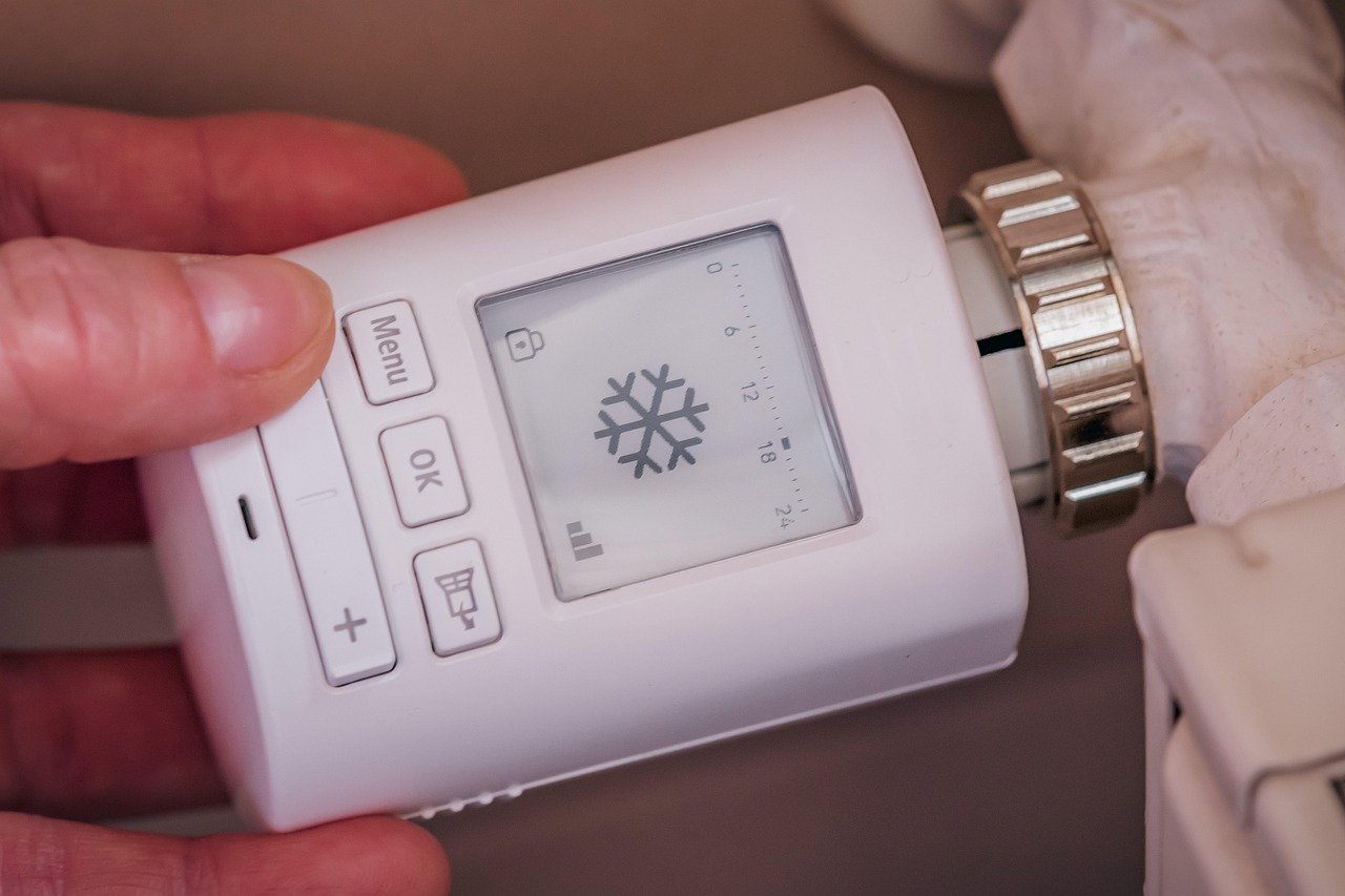 Hand holding a white handheld radiator thermostat head with a snowflake icon on its LCD and Menu, OK, and up/down buttons