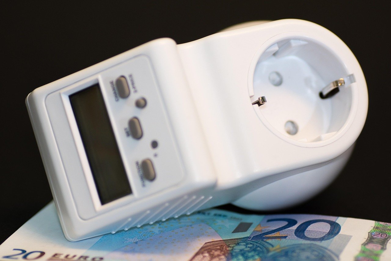 White plug-in energy meter with an LCD display, control buttons, and a socket on top, resting on two 20 euro banknotes