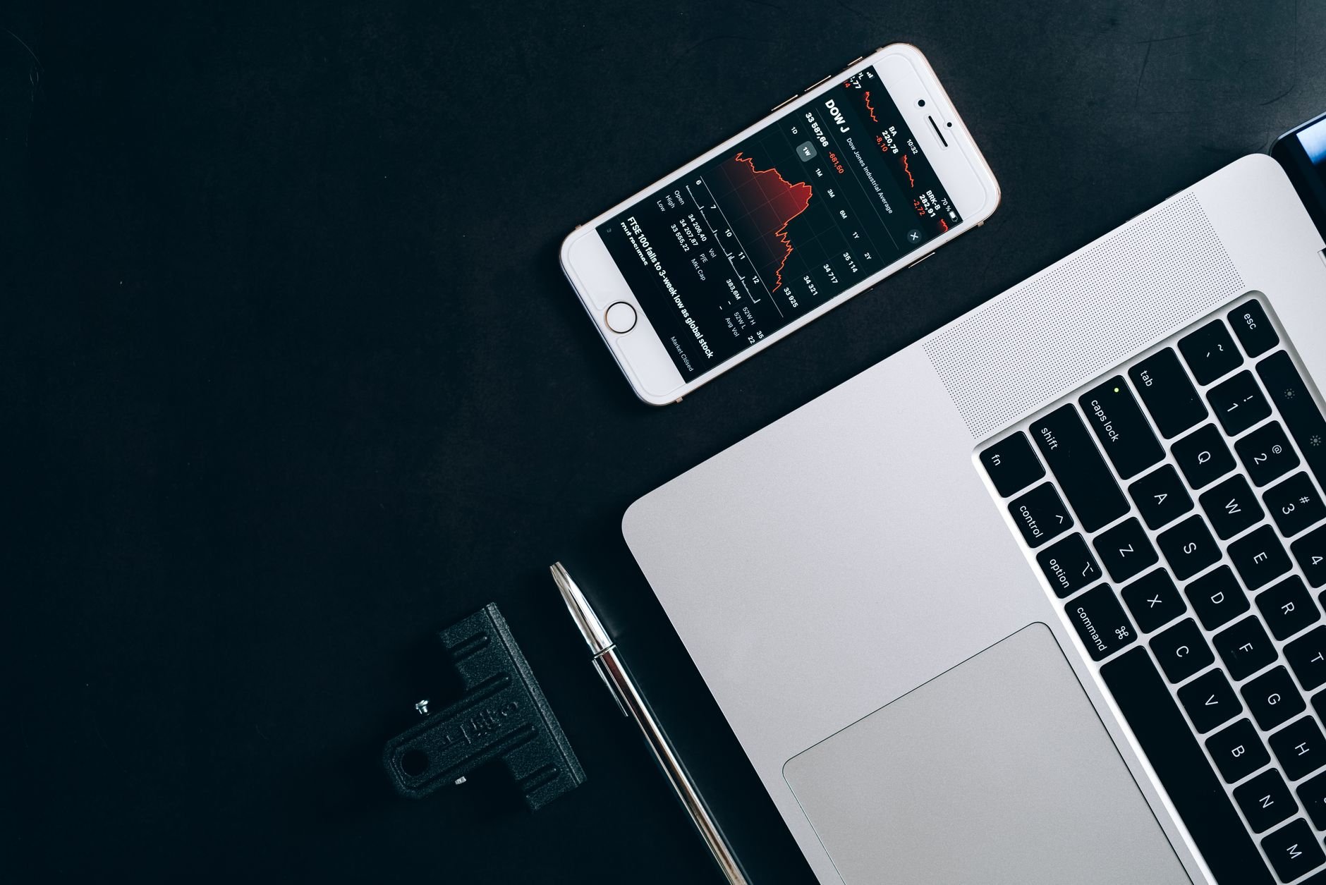 Smartphone on a dark surface beside a silver laptop and a black clip, with a red line chart on the phone screen