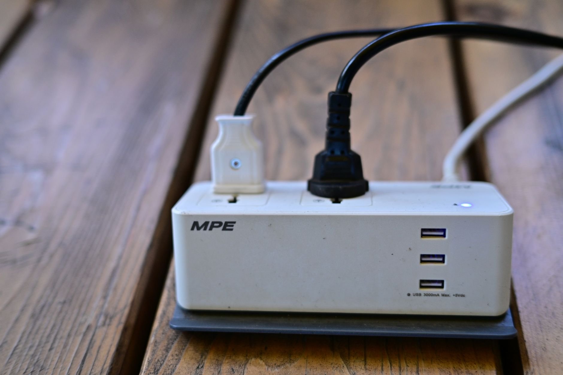 White MPE power strip with three USB ports on a wooden deck, with black and white power cords plugged into it
