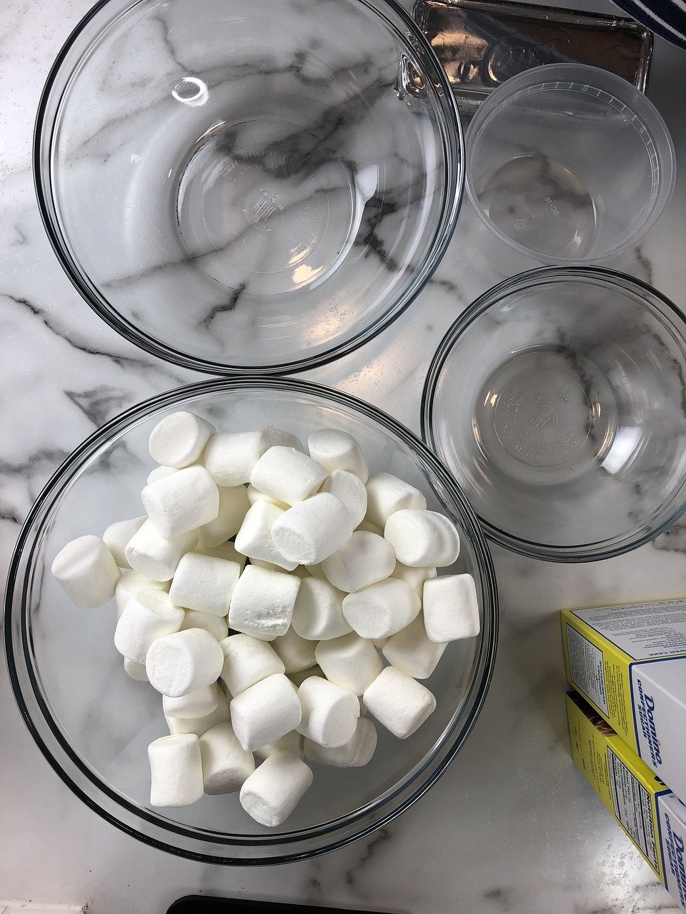 Mini marshmallows in a glass bowl beside empty glass bowls on a marble counter