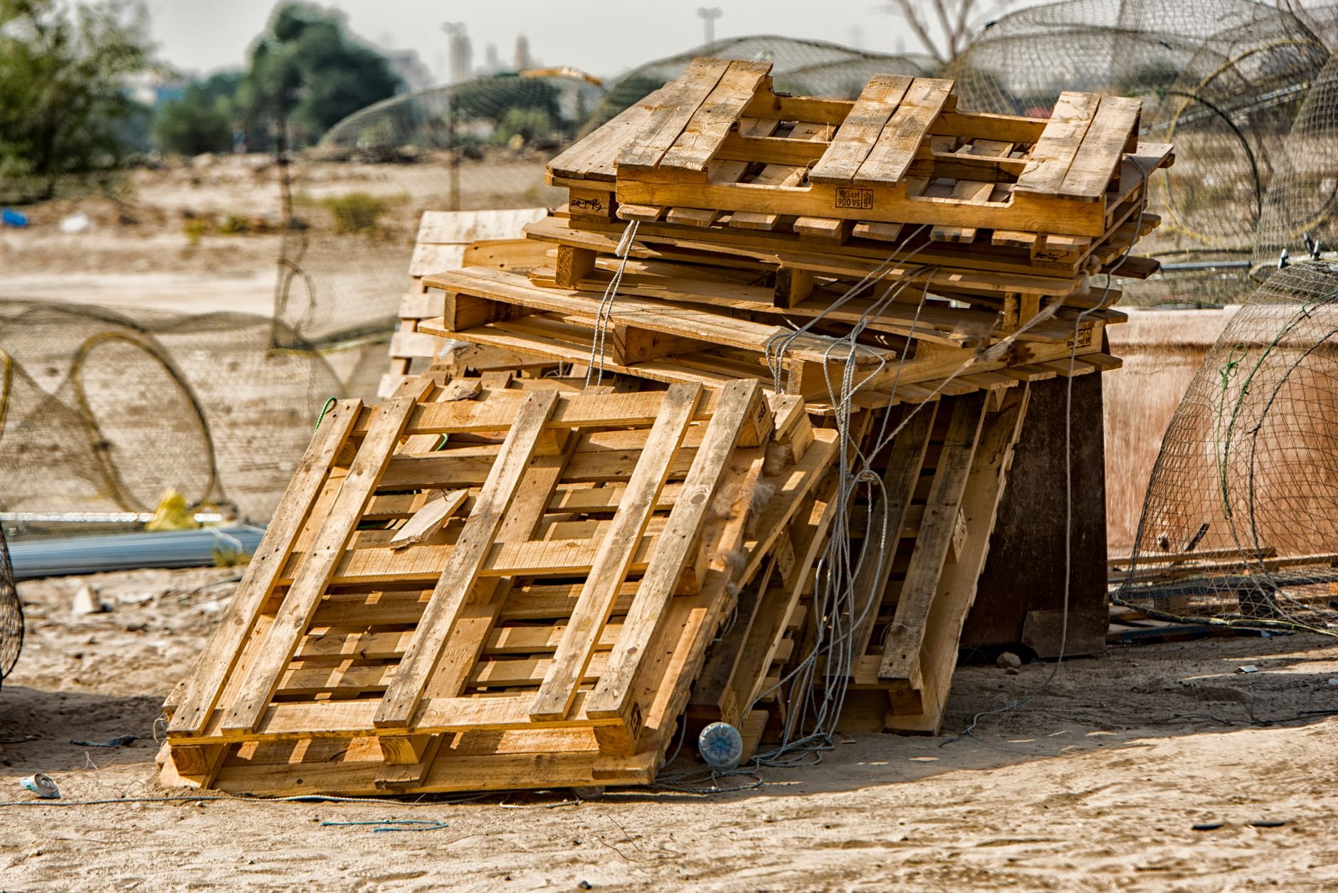 Several wooden pallets stacked outdoors on sandy ground beside metal fencing and coiled wire