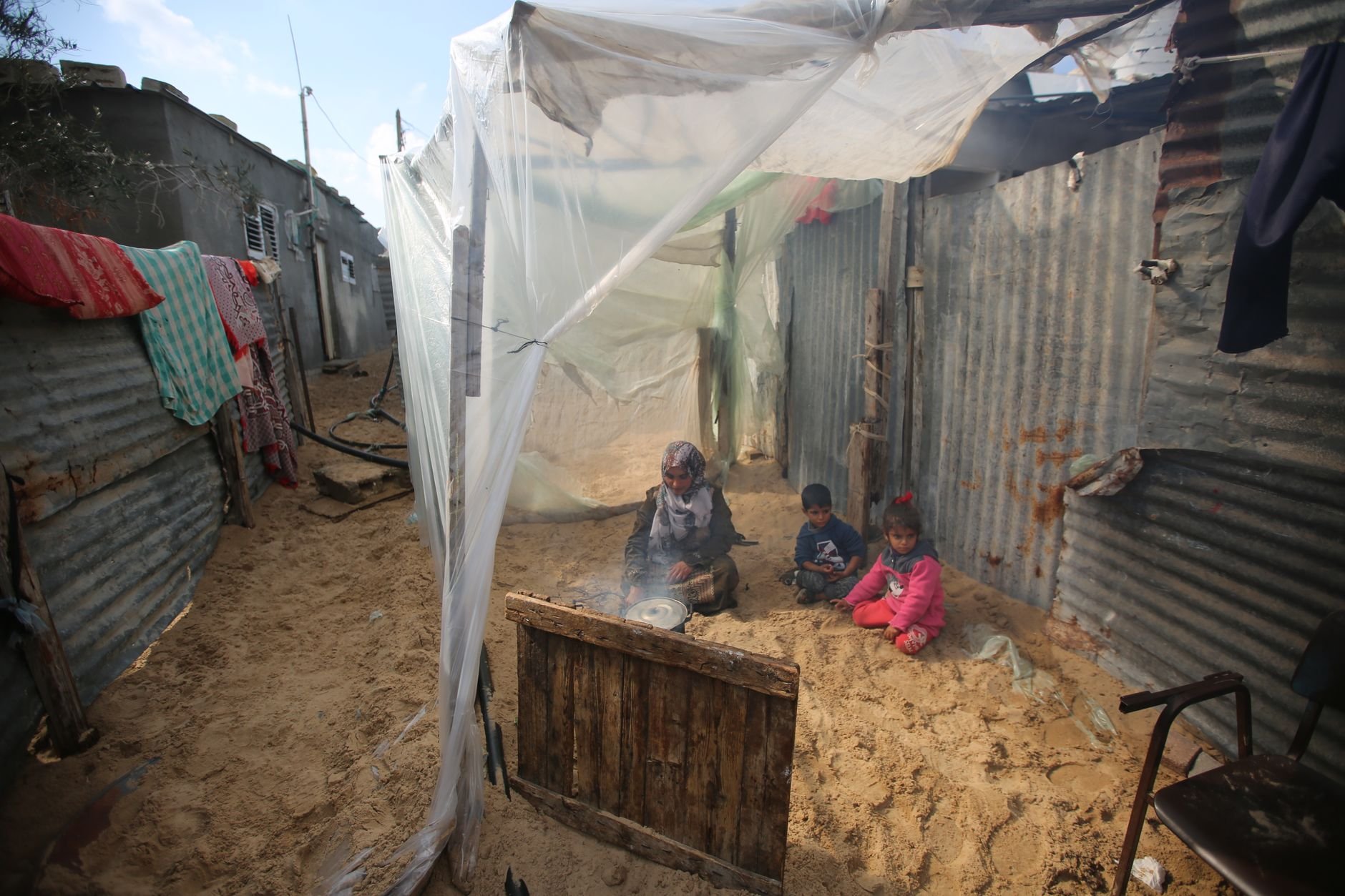 A woman and two children sit on sand inside a shelter made of plastic sheeting over a frame, with corrugated metal walls around them