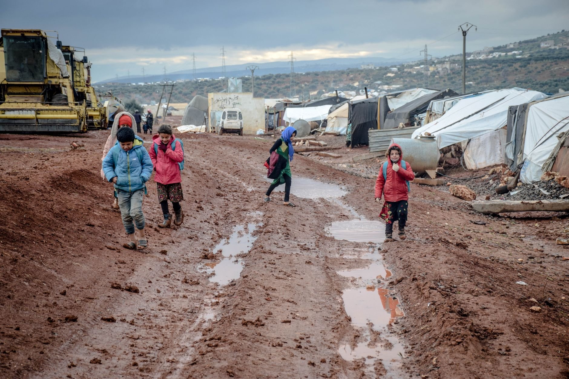 Muddy unpaved track through a camp with puddles, children walking, and tents on the right