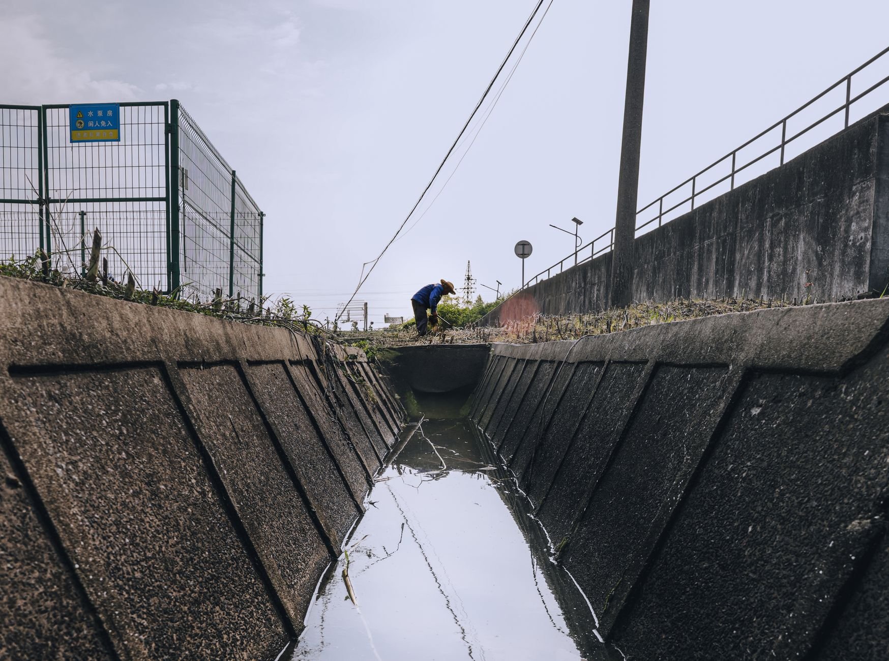 Narrow concrete drainage channel with dark water running between sloped walls, a worker bending at the far end