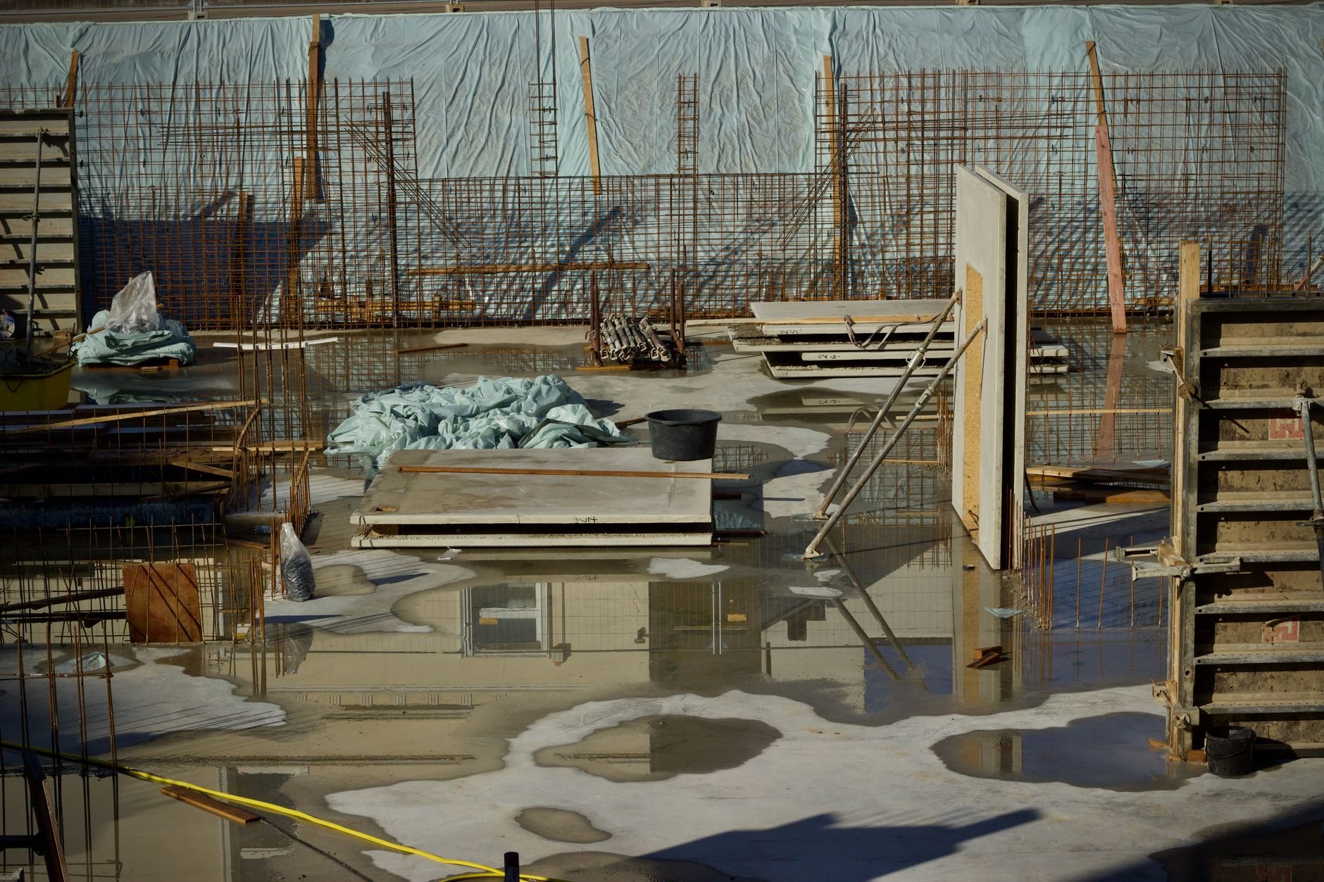 Partly flooded construction site with puddles on concrete, rebar frames, wooden boards, and a blue tarp in the background