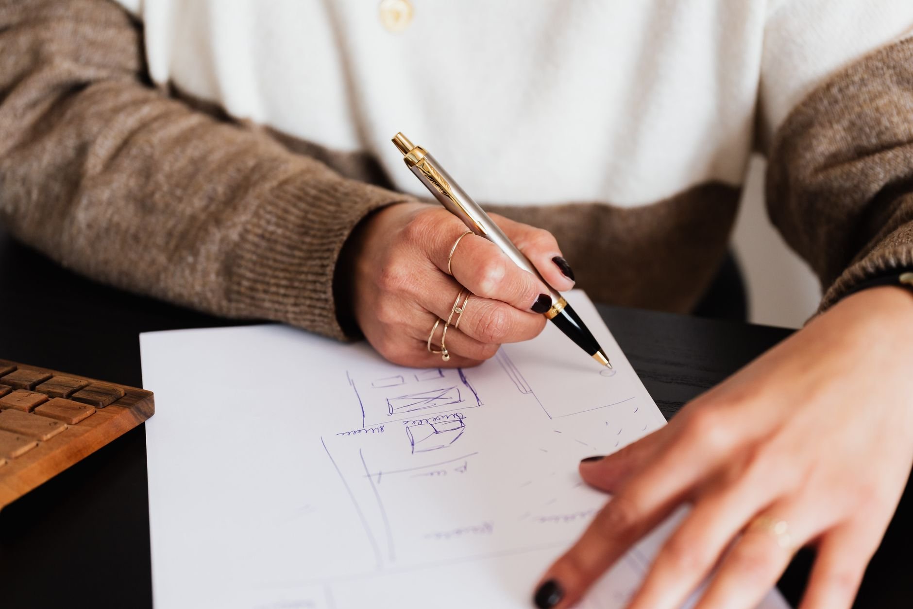 Hands holding a gold pen over a sheet of paper with a hand-sketched layout