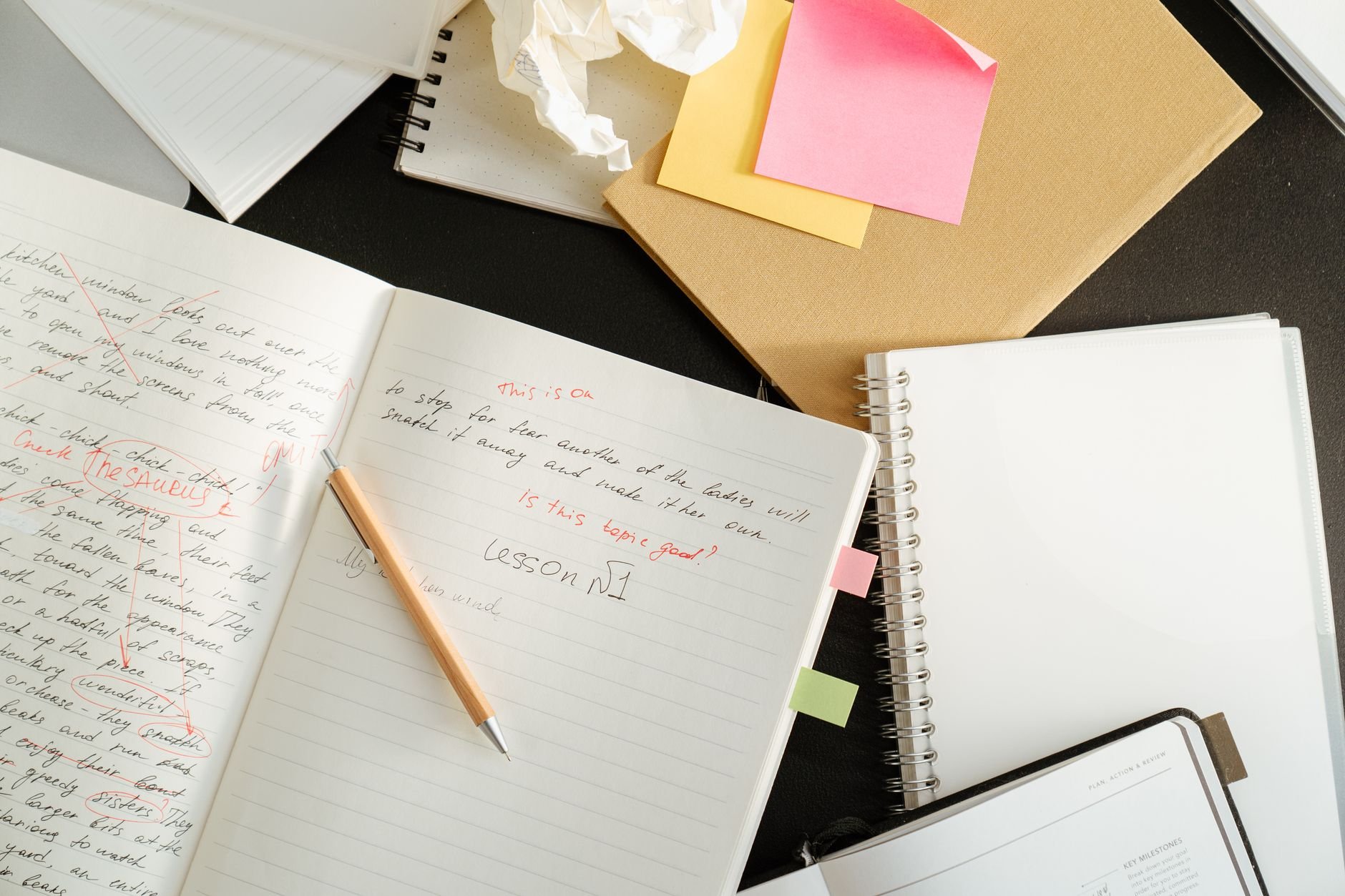 Open notebook with handwritten notes and red markings, a pencil, and sticky notes on a dark desk