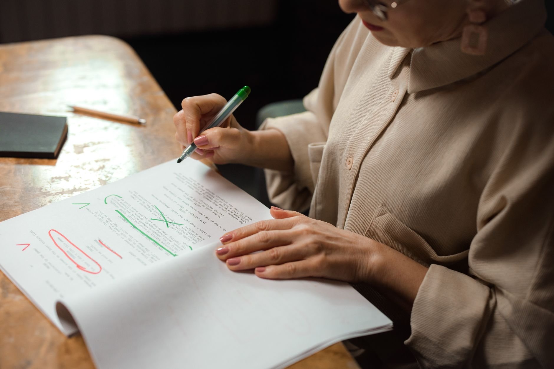 Woman in a beige blouse and glasses marking up printed pages with a green pen