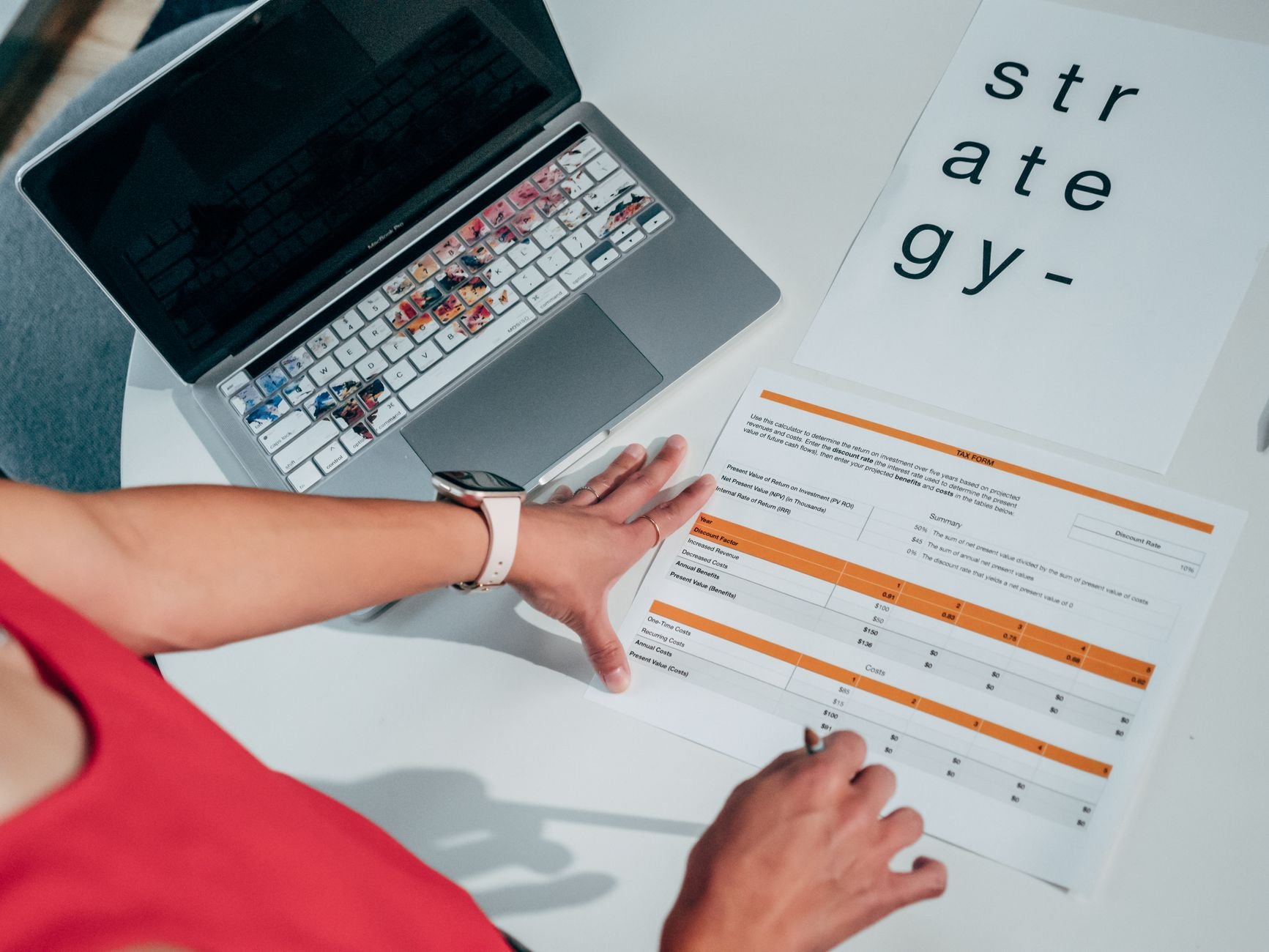 Person's hands on a white desk beside an open laptop and a printed orange-and-white table form