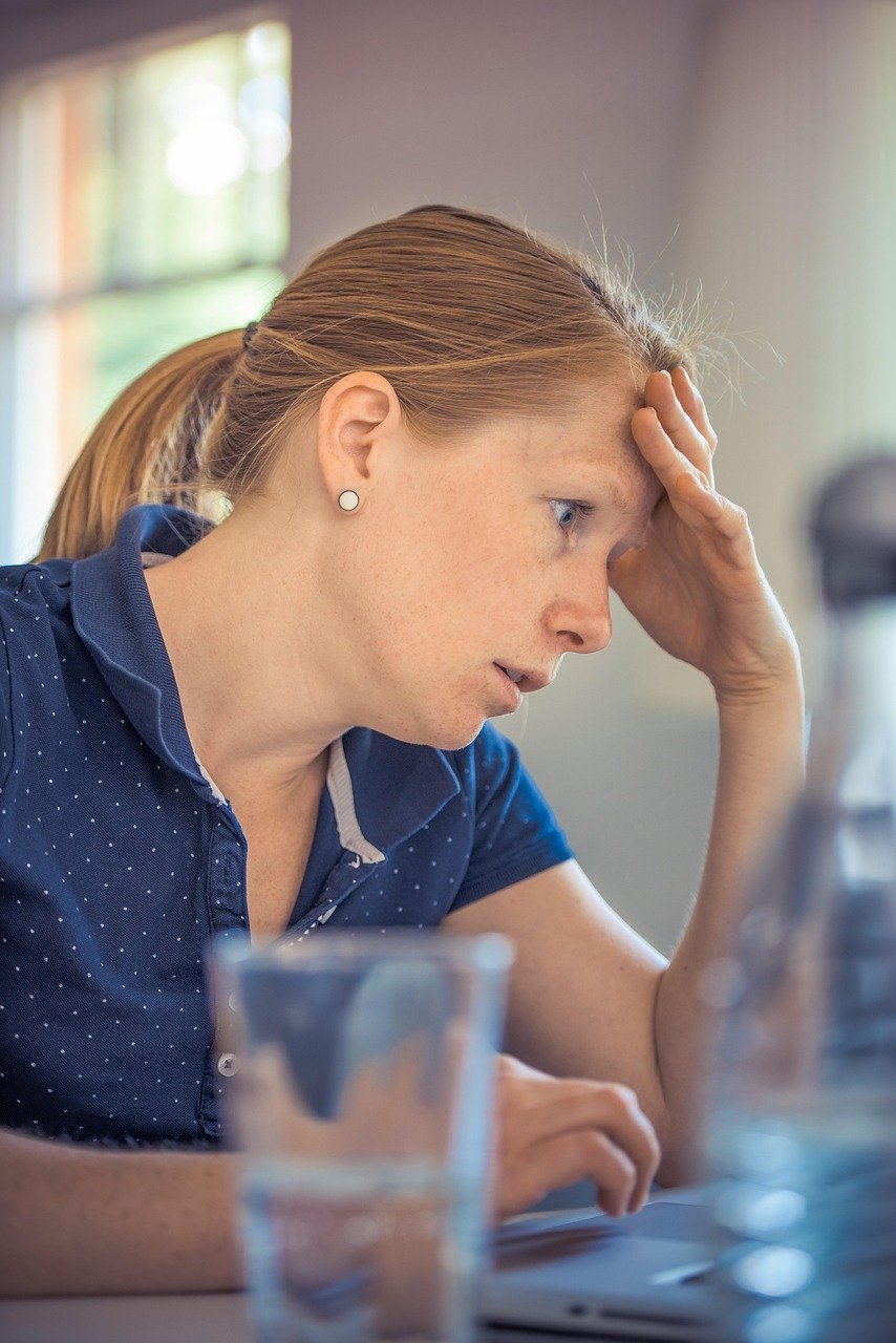 A woman with her hair pulled back rests her hand on her forehead while looking at a laptop screen, with a glass of water in the foreground