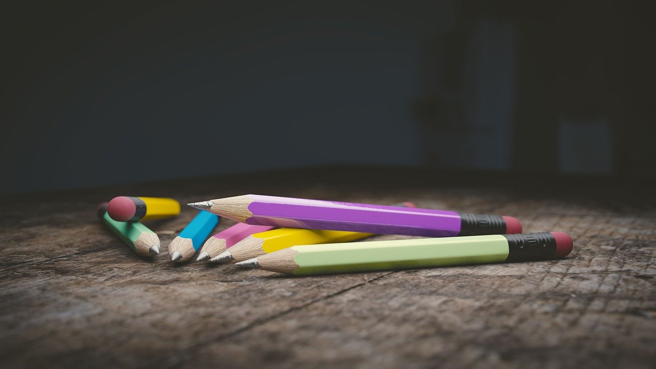 Several colored pencils with pink, purple, yellow, and green bodies and red or black erasers lying on a wooden surface