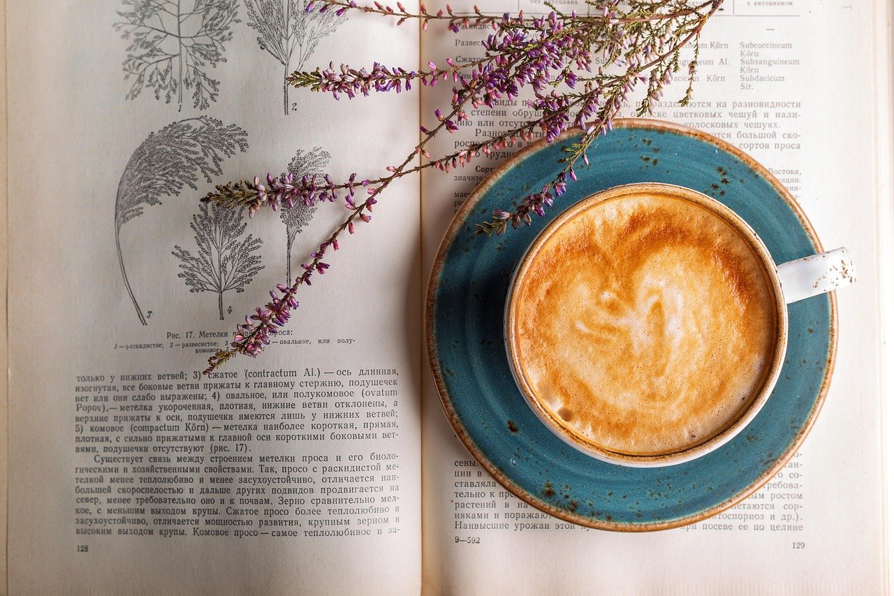 Open book with printed pages beside a cup of coffee in a blue saucer, with a sprig of purple heather lying across the page