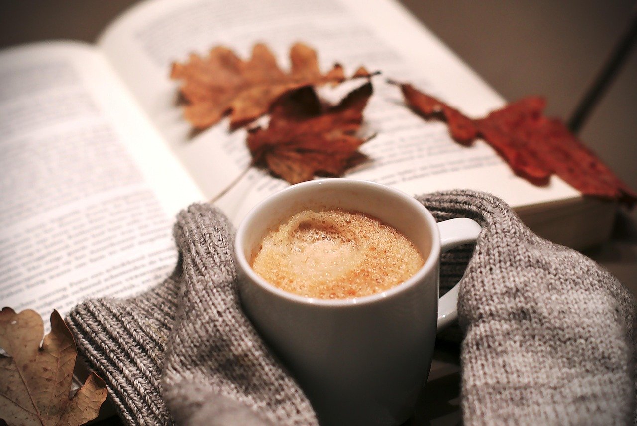 Open book with printed text pages, two dry brown autumn leaves resting on the pages, and a white cup of coffee with foam held in gray knit gloves in the foreground