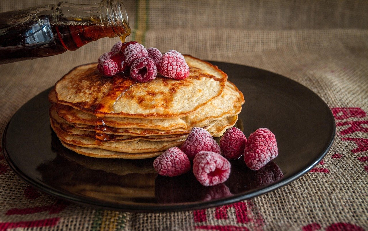 Stack of pancakes topped with frozen raspberries, with syrup being poured from a bottle onto them