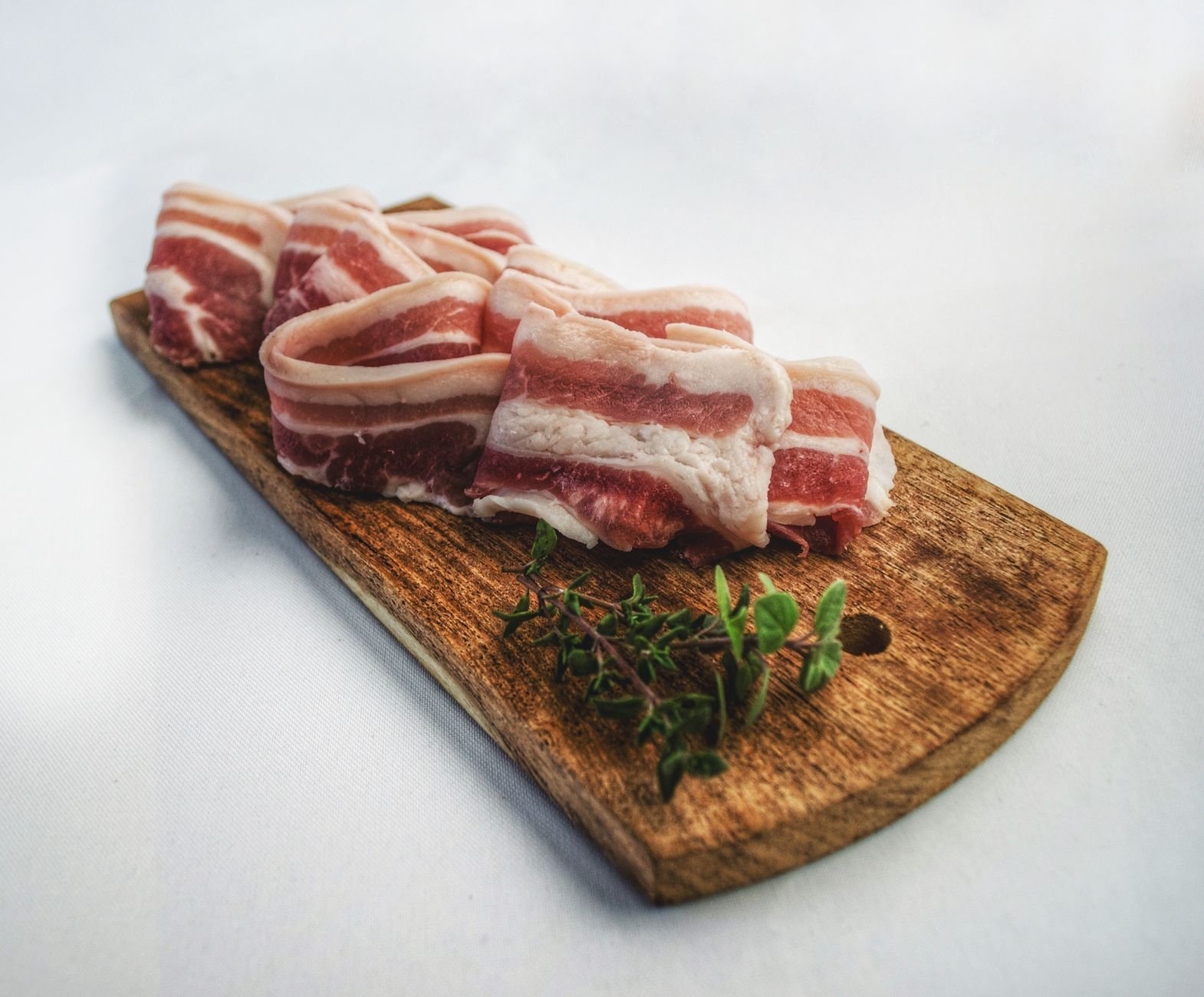 Raw bacon slices stacked on a wooden board with sprigs of fresh thyme beside them