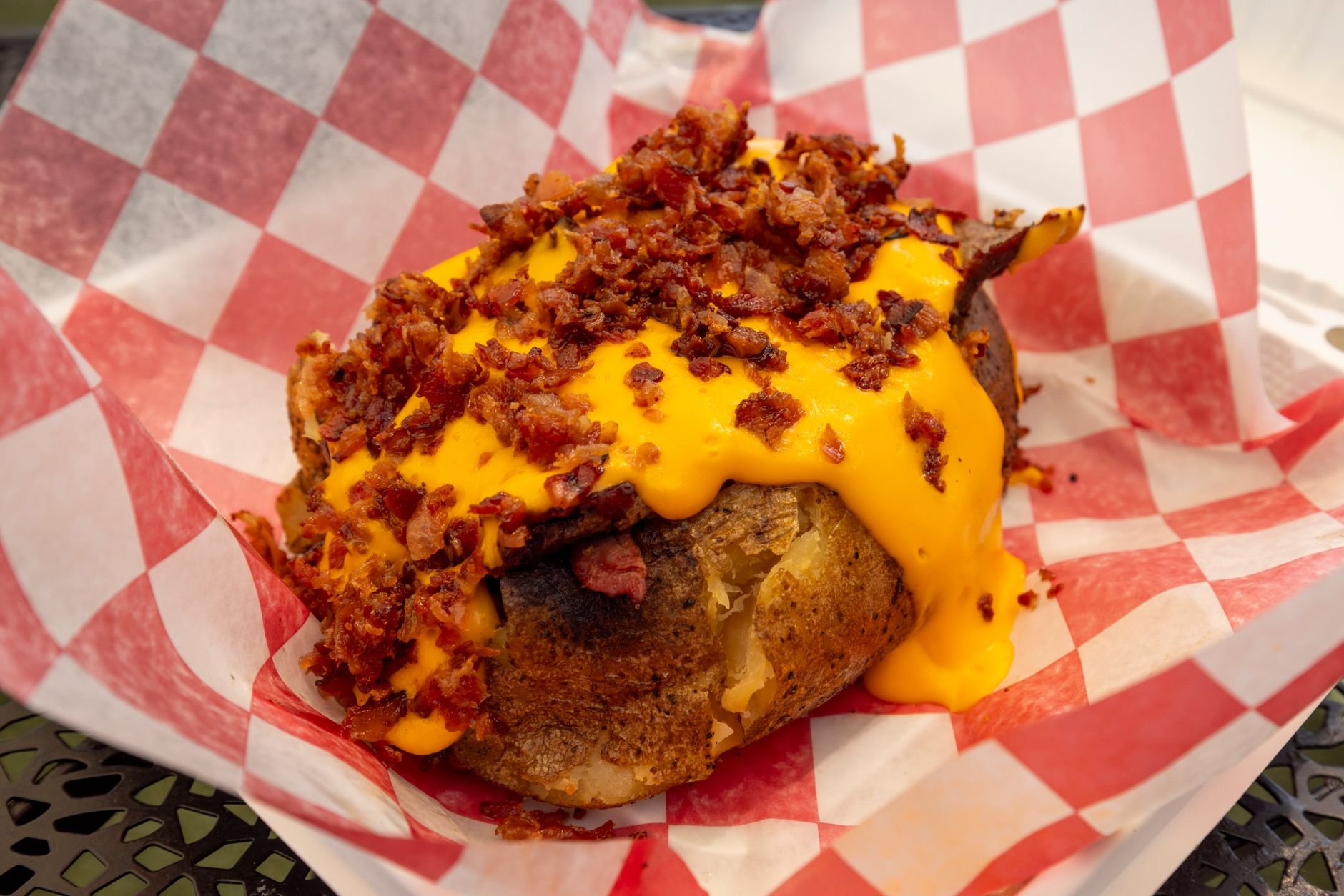 Baked potato topped with crumbled bacon and melted orange cheese sauce on a red-and-white checkered paper liner