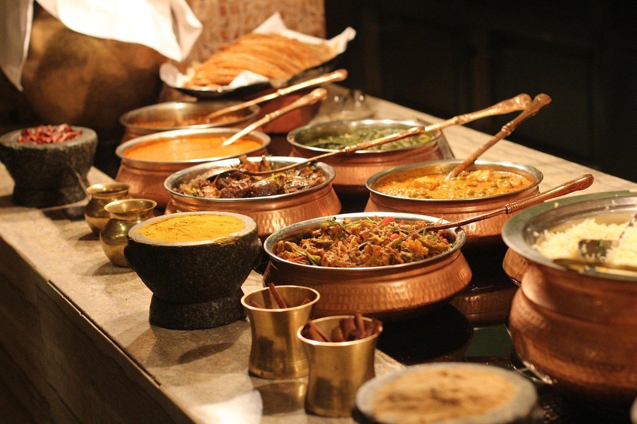 Buffet of copper pots holding curries and stews with metal serving spoons, plus brass spice cups and a bowl of yellow dip on a stone counter