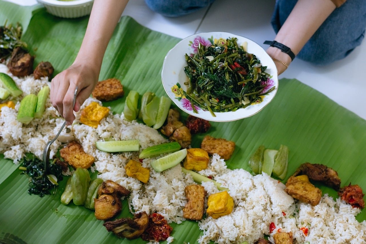 Rice served on a green banana leaf with fried pieces, sliced cucumber, vegetables and greens, while a hand holds a spoon and a plate of saut&eacute;ed greens